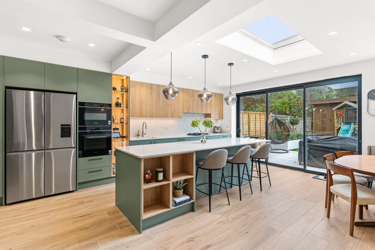 Modern kitchen design featuring an open layout with a green island, stainless steel appliances, and wooden cabinetry. The space includes stylish pendant lighting, a dining area, and large glass doors leading to an outdoor space. An Arch KBB team member is present, showcasing the inviting atmosphere of the kitchen.