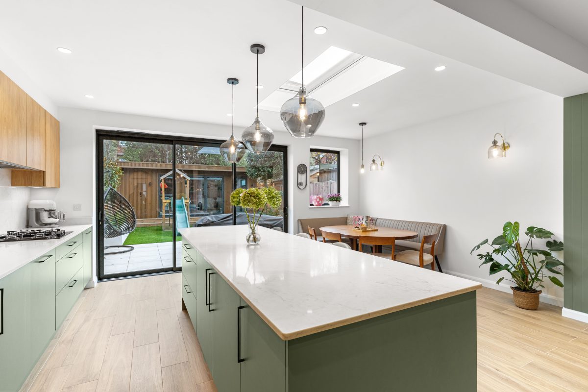 Modern kitchen interior featuring a spacious island with a marble countertop, green cabinetry, and wooden accents. The room is well-lit with pendant lighting and natural light from large sliding doors leading to a garden. An Arch KBB team member is present, and a dining area with a round wooden table and upholstered seating is visible. Lush greenery adds a touch of freshness to the space.