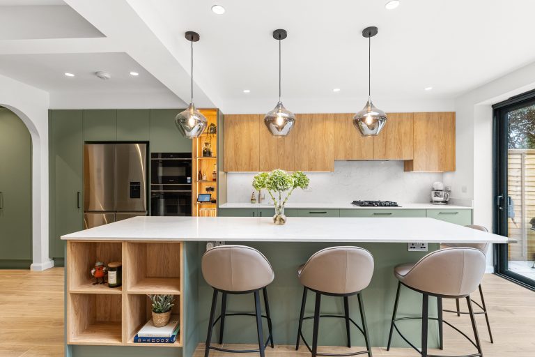 Modern kitchen featuring a large island with a white marble countertop, surrounded by stylish seating. The cabinetry combines green and wood finishes, complemented by contemporary pendant lighting. A well-equipped kitchen area includes a refrigerator and built-in appliances, creating a functional and inviting space. An Arch KBB team member is present, enhancing the atmosphere of the kitchen design.