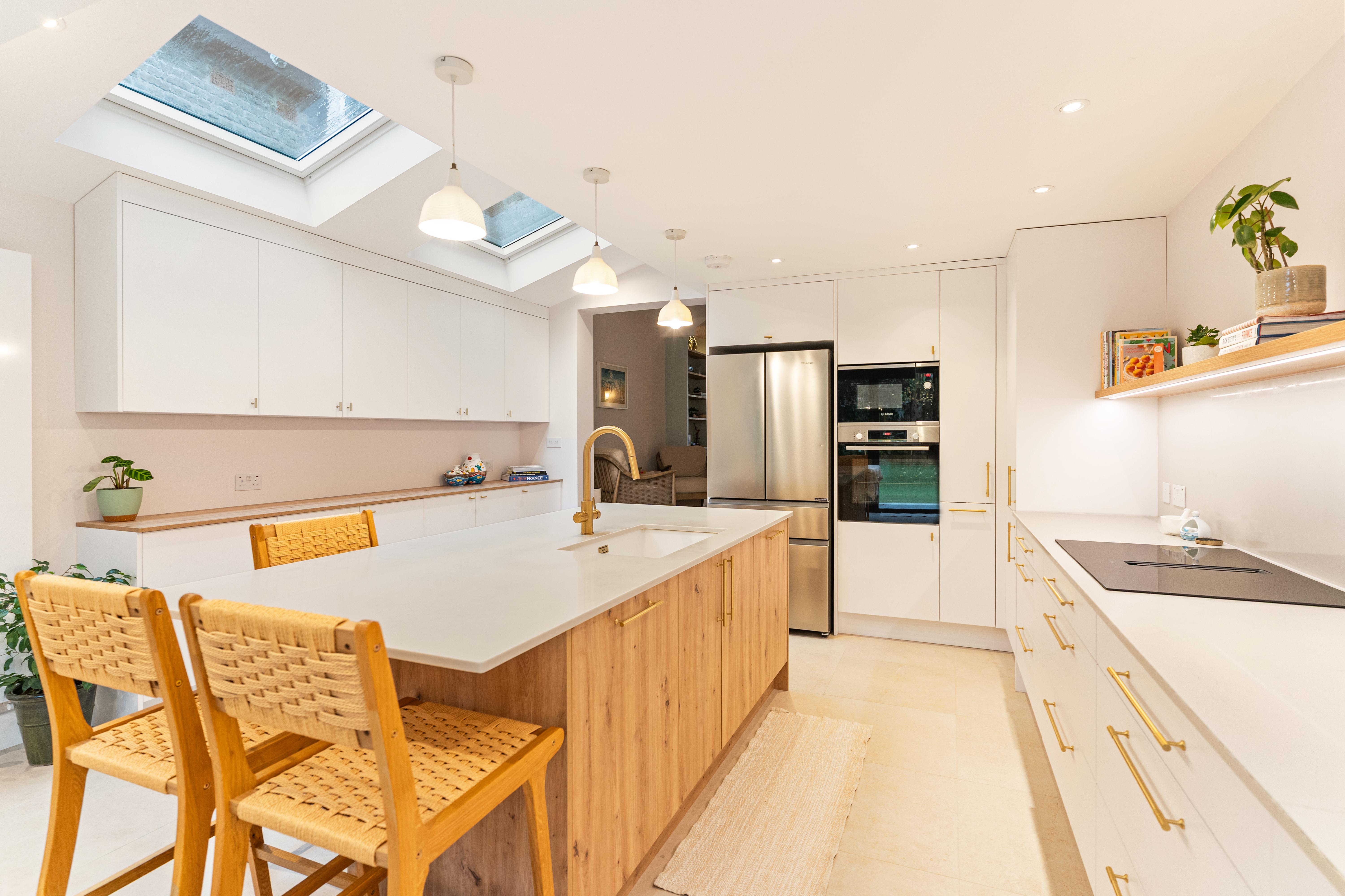 Modern kitchen featuring a spacious island with a wooden base and white countertop, two woven bar stools, sleek cabinetry, integrated appliances, and natural light streaming through skylights. An Arch KBB team member is present, enhancing the design experience.