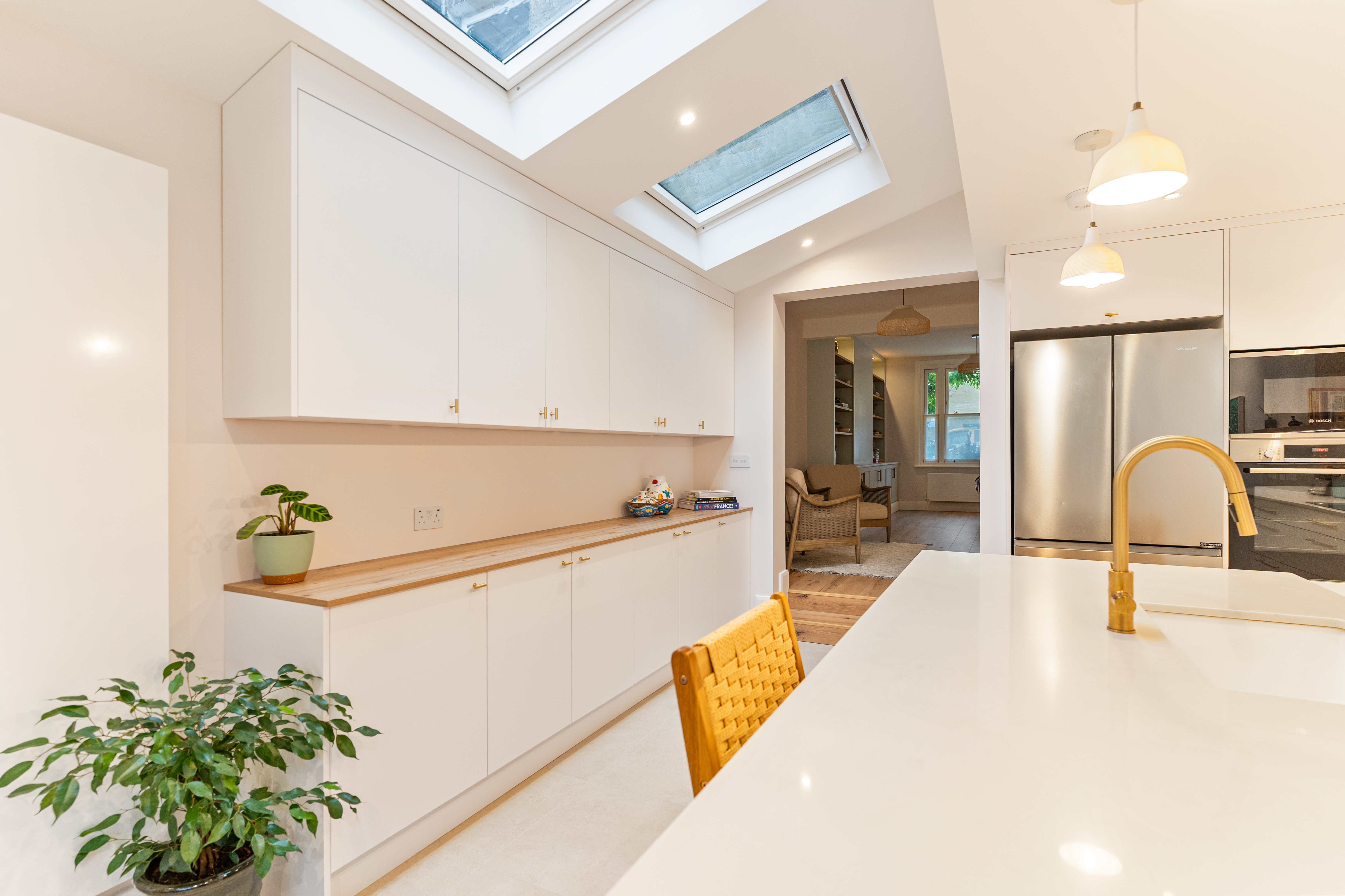 Modern kitchen interior featuring white cabinetry, a wooden countertop, and a potted plant. Skylights provide natural light, enhancing the space's ambiance. An Arch KBB team member is present, contributing to the kitchen's design. The background includes a living area with a comfortable chair and shelving.