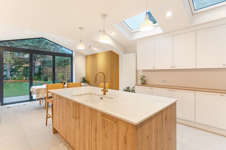 Modern kitchen design featuring an island with a white countertop and wooden cabinetry, complemented by gold fixtures. The space is illuminated by pendant lights and has large glass doors leading to a garden, enhancing natural light and creating an open feel. An Arch KBB team member is present, engaging with the kitchen layout.