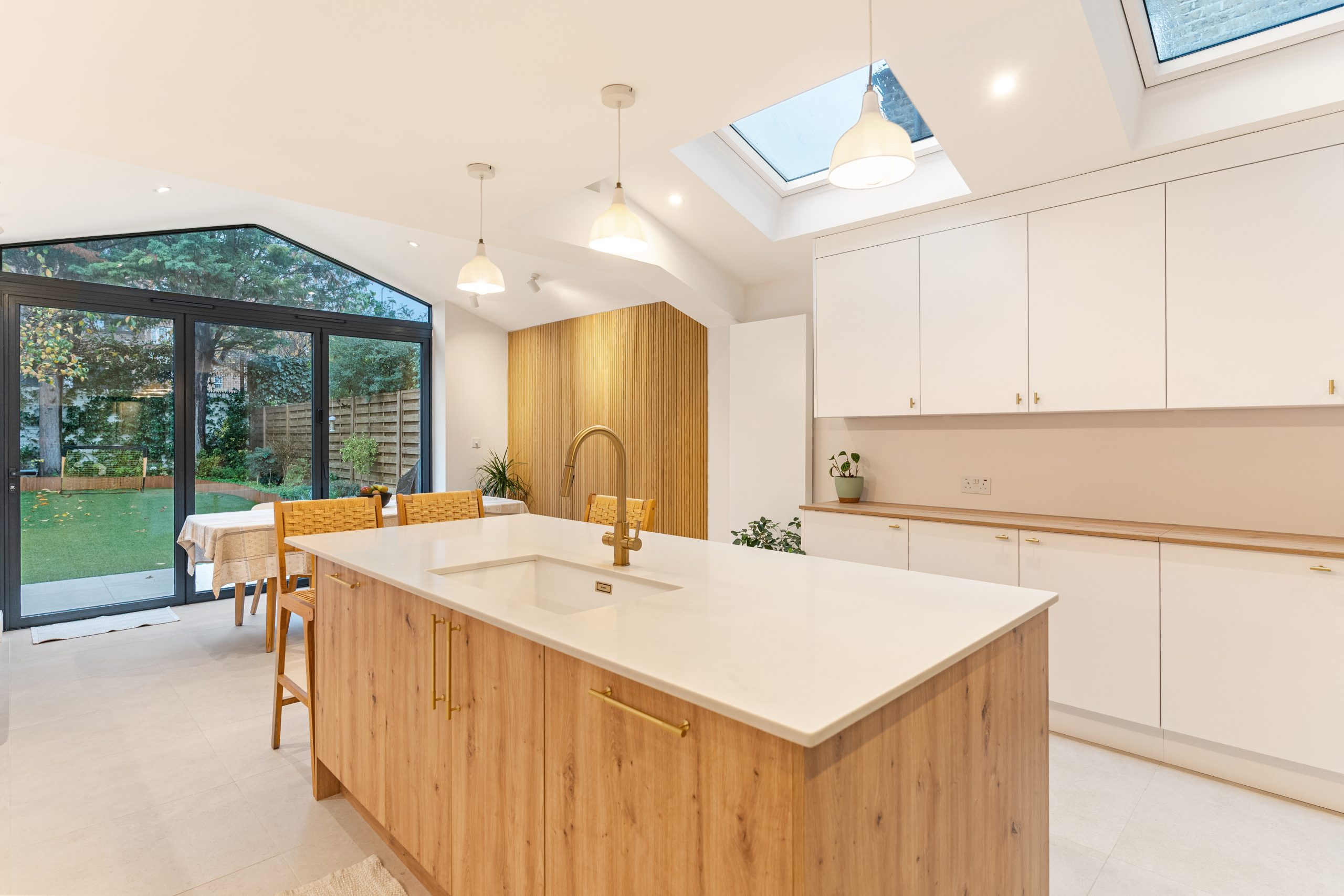 Modern kitchen design featuring an island with a white countertop and wooden cabinetry, complemented by gold fixtures. The space is illuminated by pendant lights and has large glass doors leading to a garden, enhancing natural light and creating an open feel. An Arch KBB team member is present, engaging with the kitchen layout.