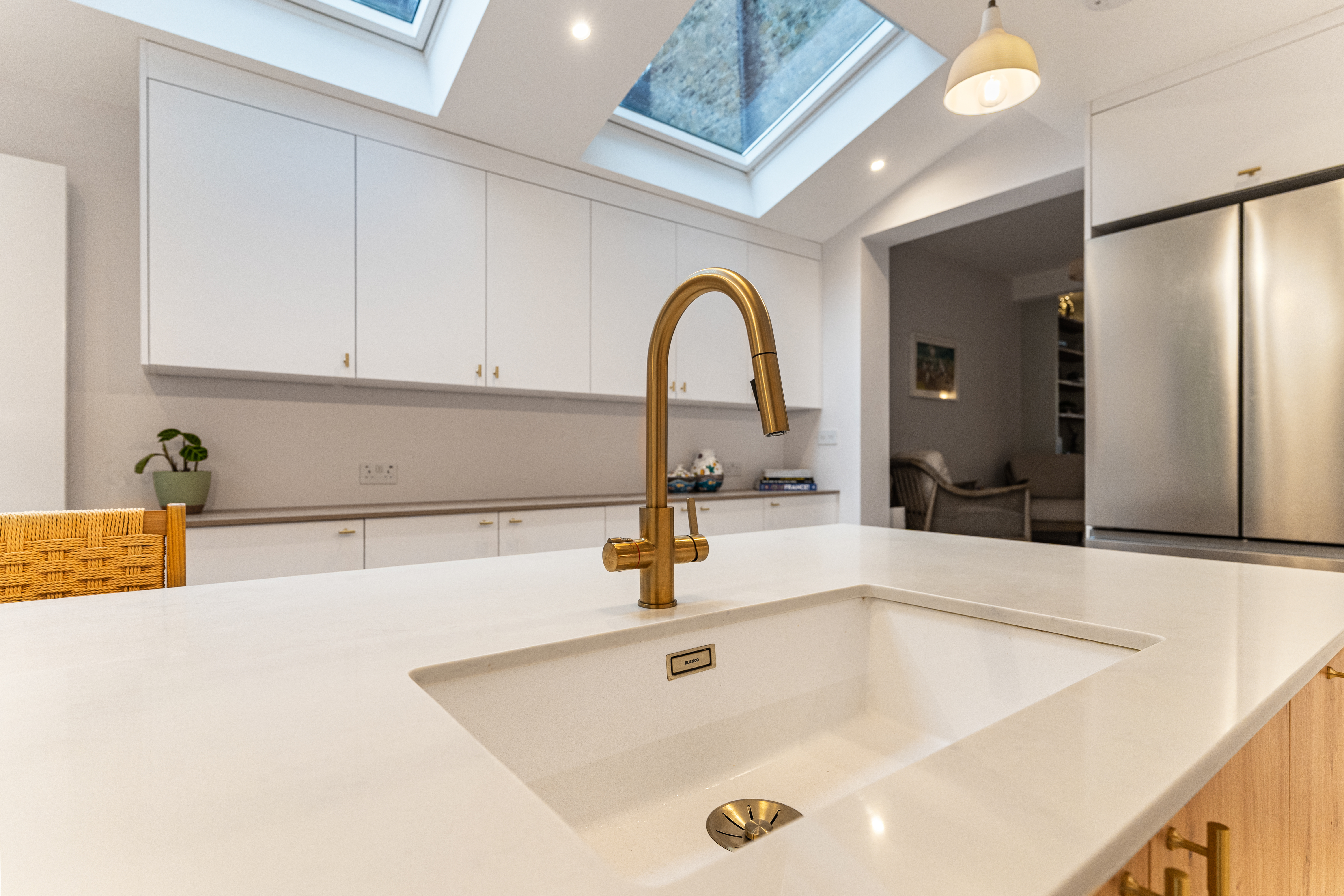 Modern kitchen featuring a sleek white countertop and a stylish gold sink tap, with ample storage cabinets in the background. An Arch KBB team member is visible, showcasing the functional design of the space, illuminated by natural light from the skylights above.