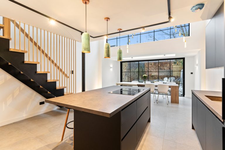 Modern kitchen interior featuring a stylish island with a built-in hob, pendant lighting, and a staircase with wooden and black accents. Large windows provide natural light, illuminating the dining area with a wooden table and white chairs. An Arch KBB team member is present, showcasing the space's design.