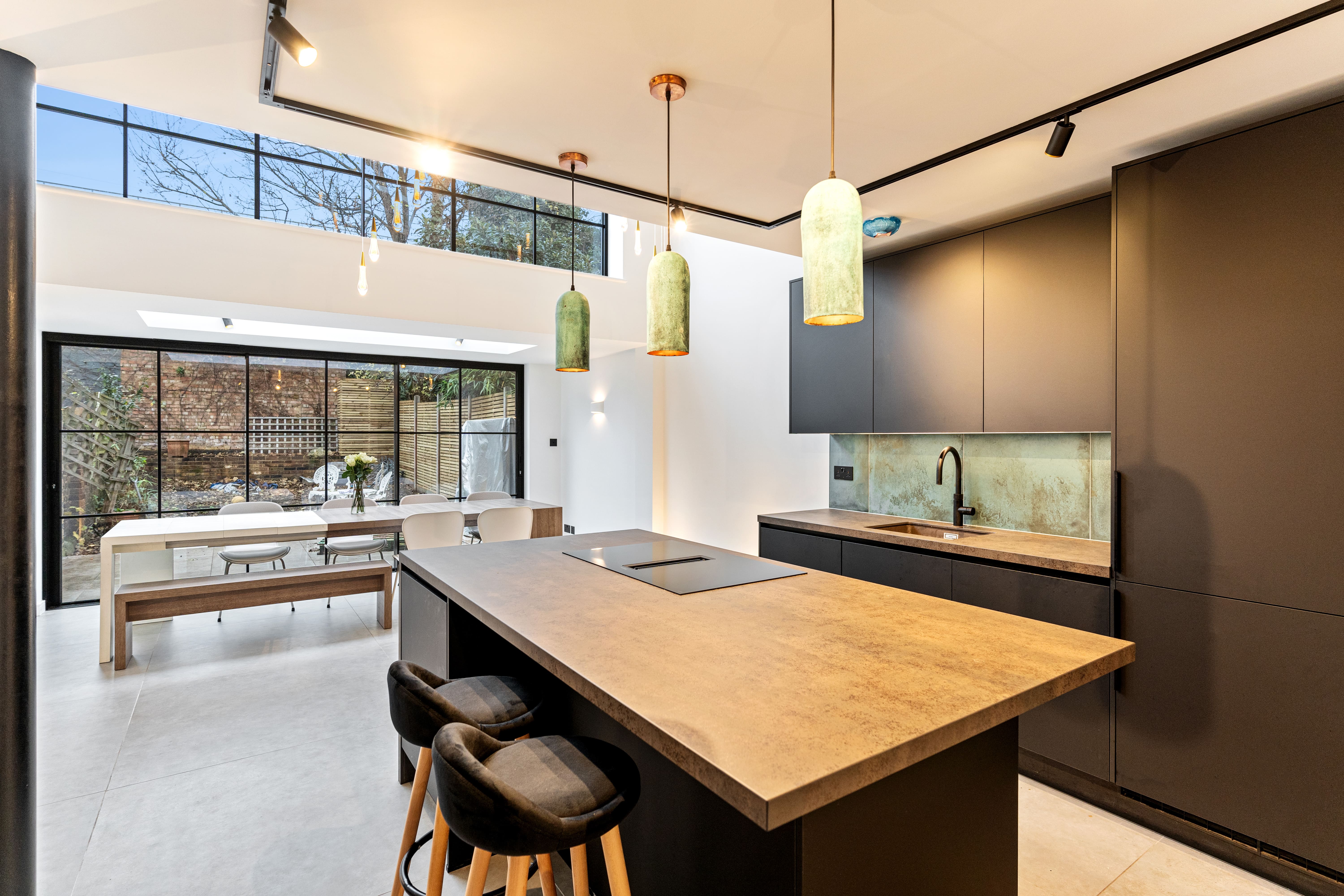 Modern kitchen interior featuring a sleek island with a textured countertop, black cabinetry, and stylish pendant lighting. Large windows allow natural light to flood the space, highlighting a dining area with a wooden table and white chairs. An Arch KBB team member is present, showcasing the contemporary design and functionality of the kitchen.