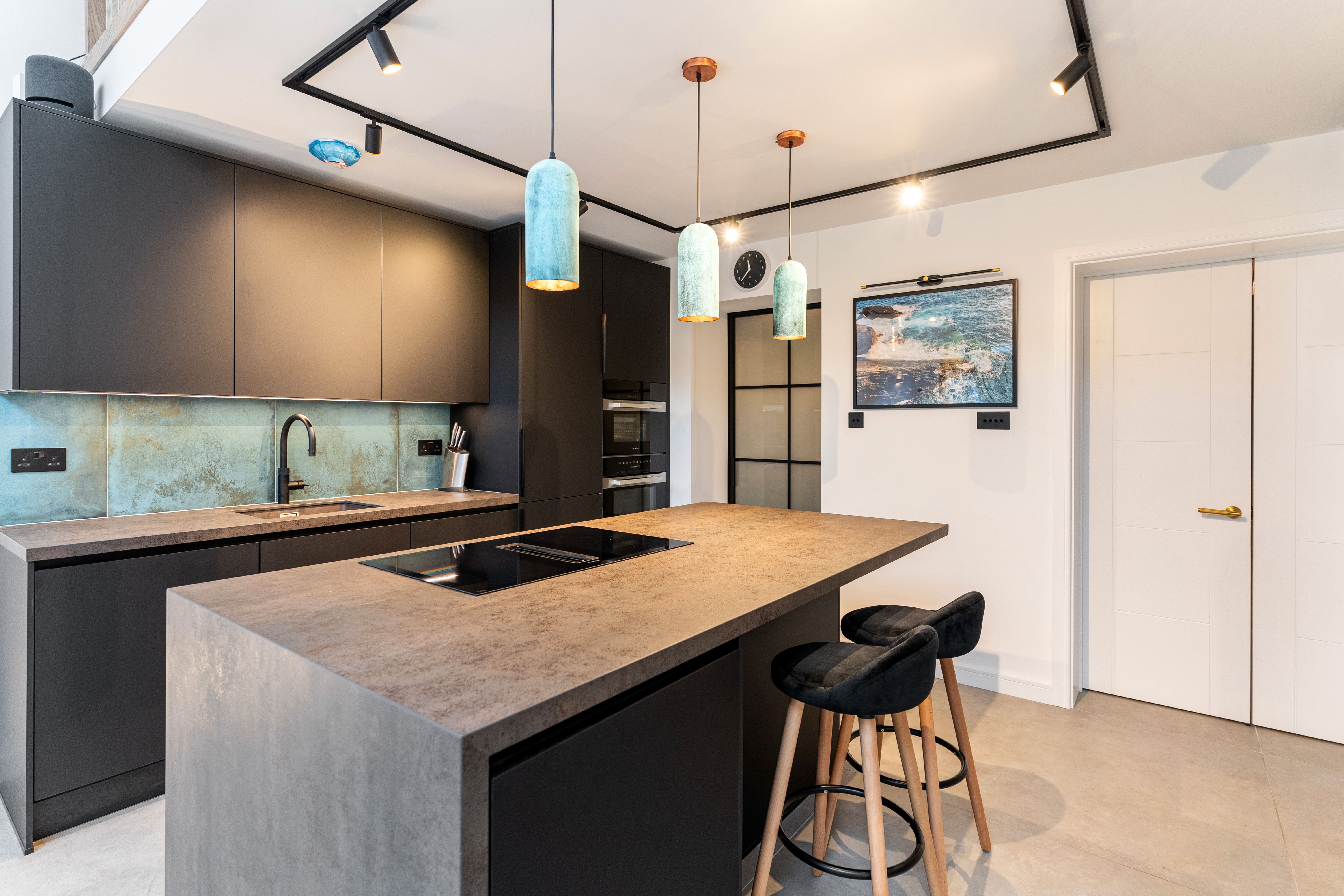 Modern kitchen interior featuring sleek black cabinetry, a concrete countertop, and stylish pendant lighting. The design includes a blue-tinted splashback and integrated appliances, creating a contemporary aesthetic. An Arch KBB team member is present, showcasing the space's functionality and elegance.