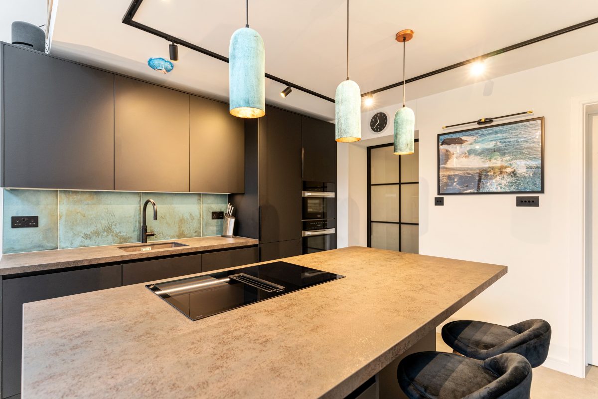 Modern kitchen design featuring sleek black cabinetry, a textured stone countertop, and stylish pendant lighting. An Arch KBB team member is present, showcasing the contemporary layout that includes a built-in oven and an integrated sink. The blue-green backsplash adds a pop of colour, complemented by a wall-mounted clock and framed artwork.