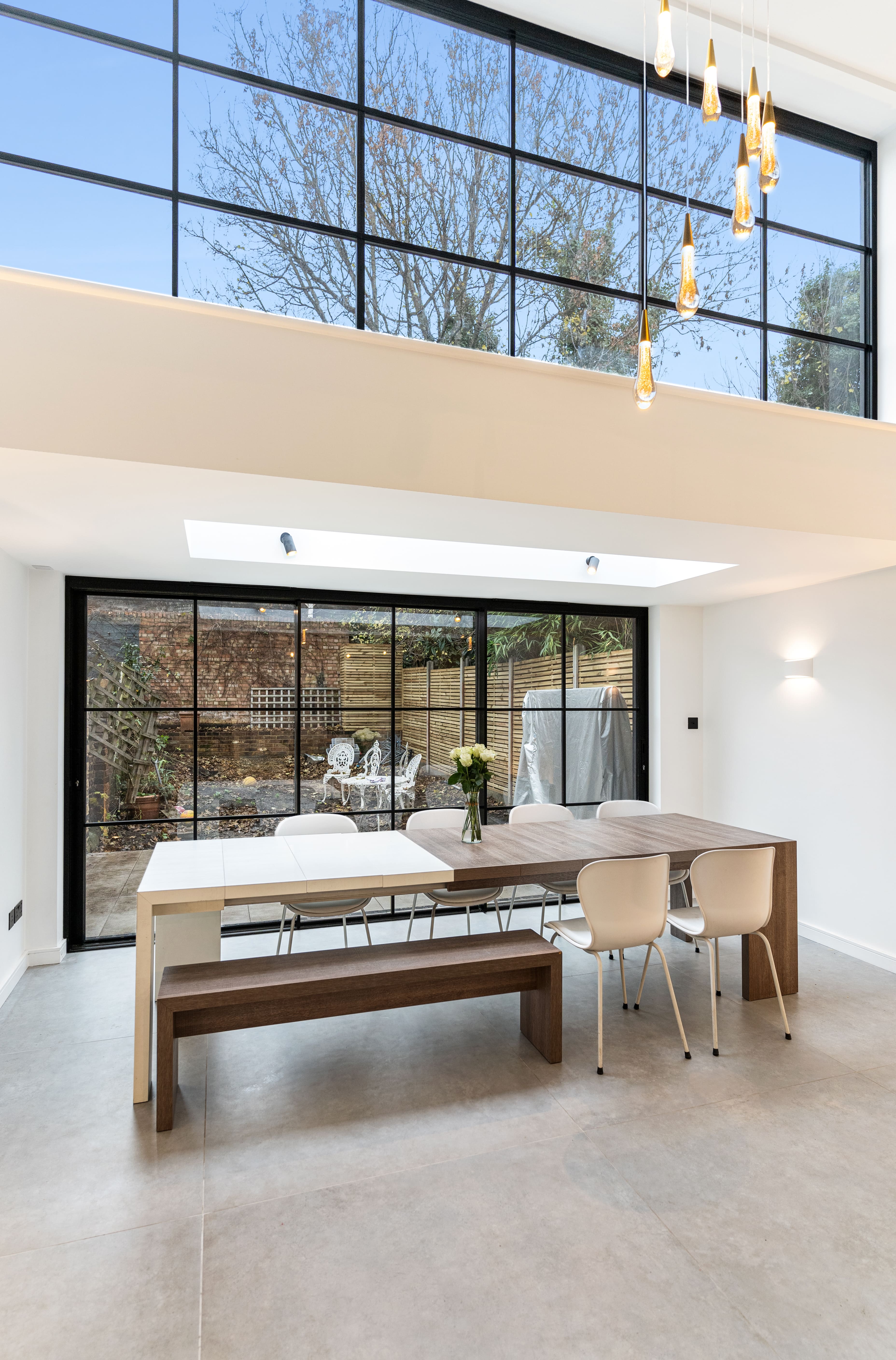 Bright and modern dining area featuring a large wooden table with white chairs, complemented by a stylish bench. Large floor-to-ceiling windows provide ample natural light and a view of the garden, enhancing the open space. An Arch KBB team member is present, showcasing the contemporary interior design.