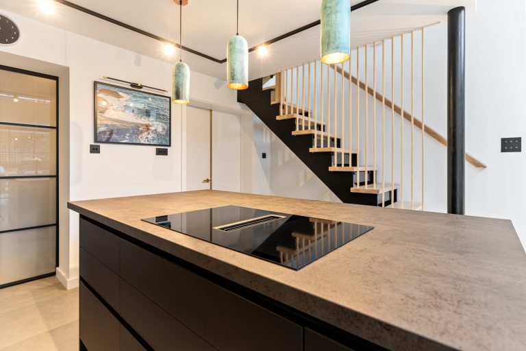 Modern kitchen interior with a sleek black induction hob on a central island, pendant lighting, and a stylish open staircase in the background.