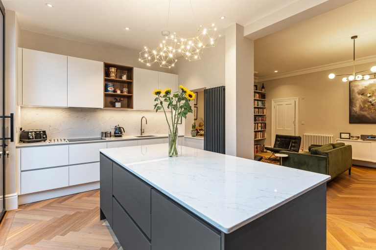 Modern kitchen interior featuring a marble island, sleek cabinetry, and stylish lighting fixtures. A vase of sunflowers adds a touch of colour, while an Arch KBB team member engages with the space. The layout includes a dining area and bookshelf, showcasing a blend of contemporary design and functionality.