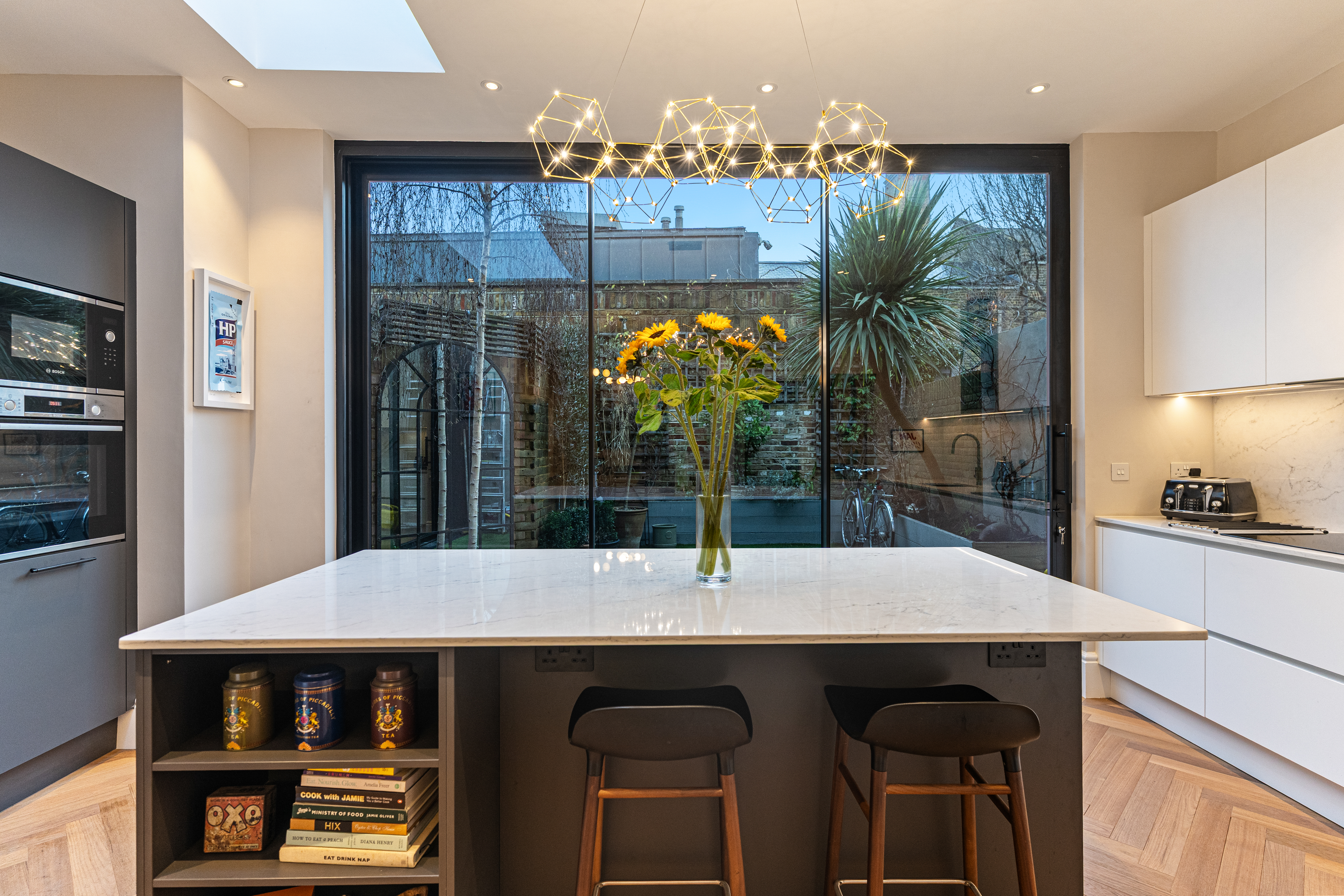 Modern kitchen interior featuring a large marble island with seating, a vase of sunflowers, and sleek cabinetry. Large glass doors provide a view of a landscaped garden, enhancing natural light in the space. An Arch KBB team member is present, showcasing the kitchen's design.
