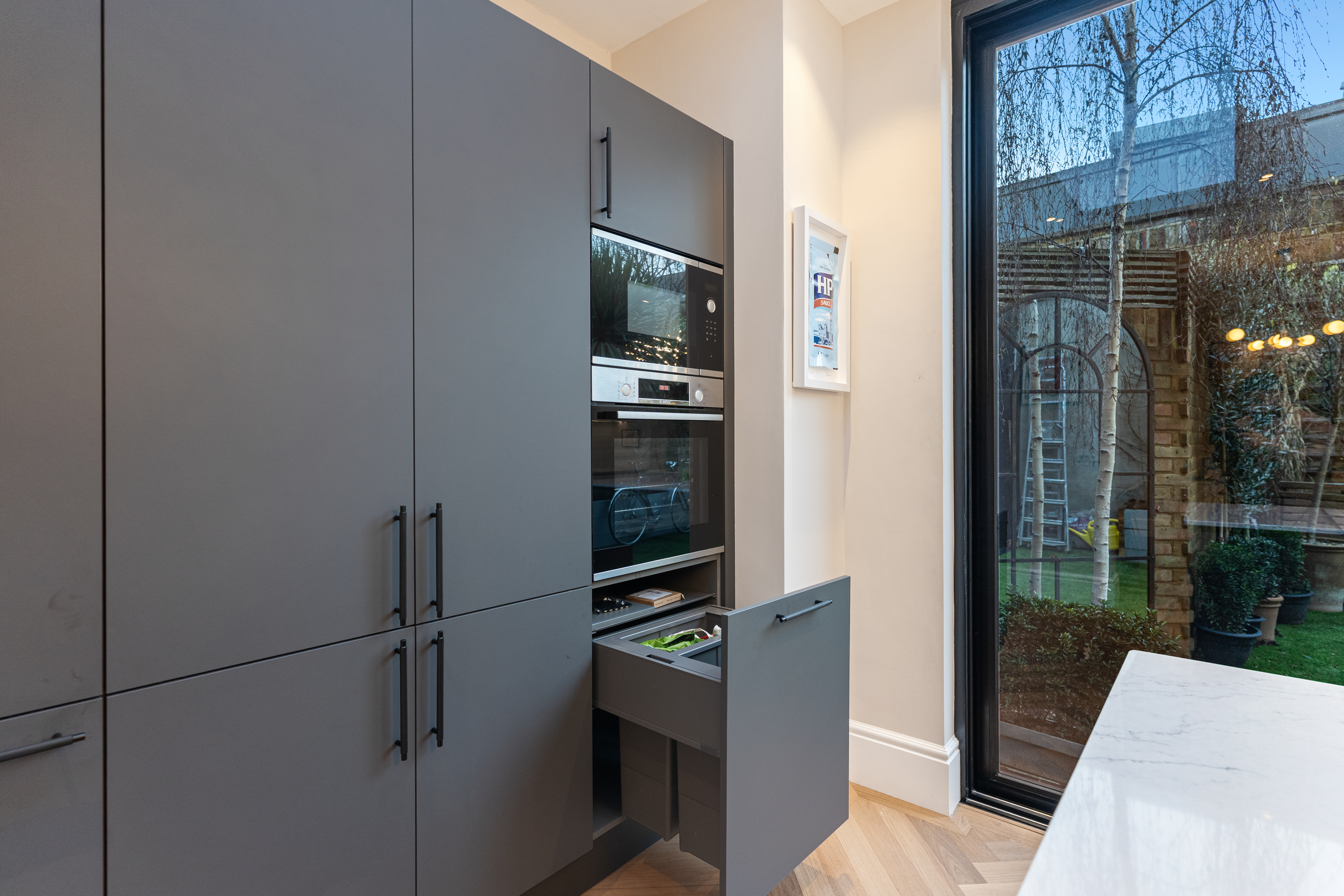 Modern kitchen featuring sleek grey cabinetry with an open drawer displaying utensils. Built-in appliances, including an oven and microwave, are seamlessly integrated. A large window offers a view of a garden area, enhancing the space's natural light. An Arch KBB team member is present, contributing to the kitchen's design and functionality.