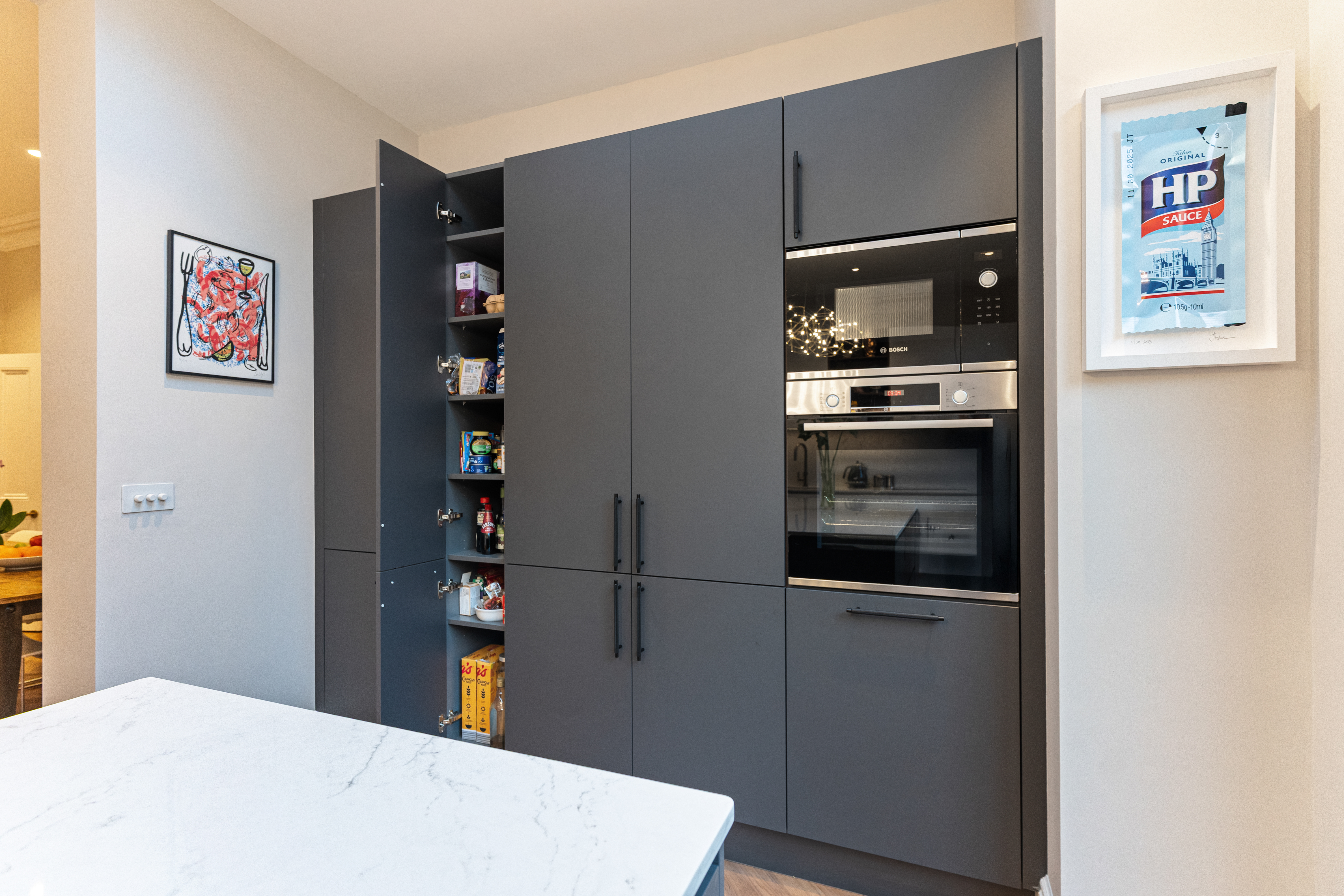 Modern kitchen featuring sleek grey cabinetry, built-in Bosch appliances, and a stylish marble countertop. An Arch KBB team member is present, showcasing the functional design and storage solutions. A piece of artwork and a framed sauce packet add character to the space.
