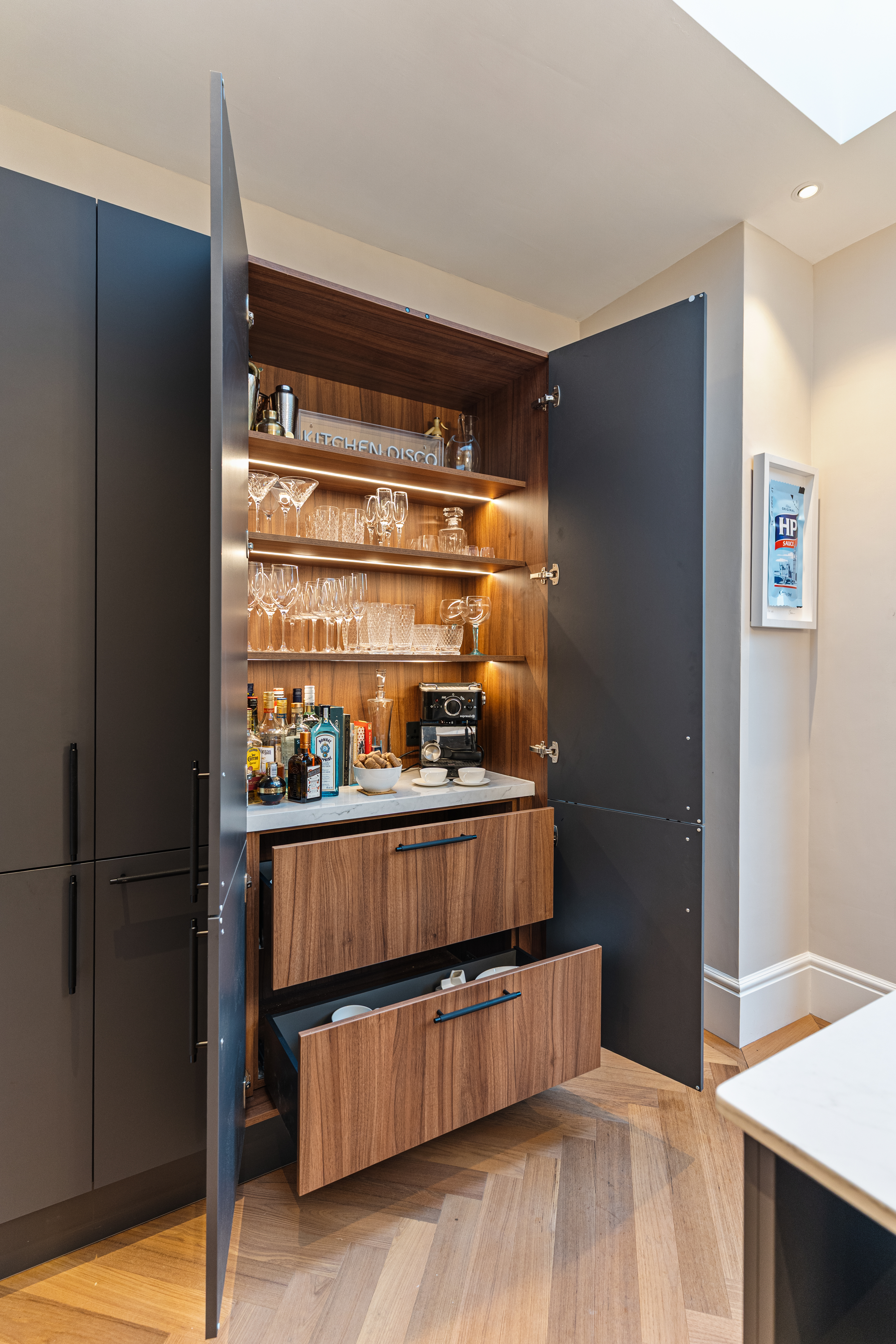 A stylish kitchen cabinet with open doors revealing shelves stocked with glassware, a coffee machine, and various bottles. The warm wooden finish and modern design elements create an elegant storage solution. An Arch KBB team member is present, showcasing the functionality of the space.