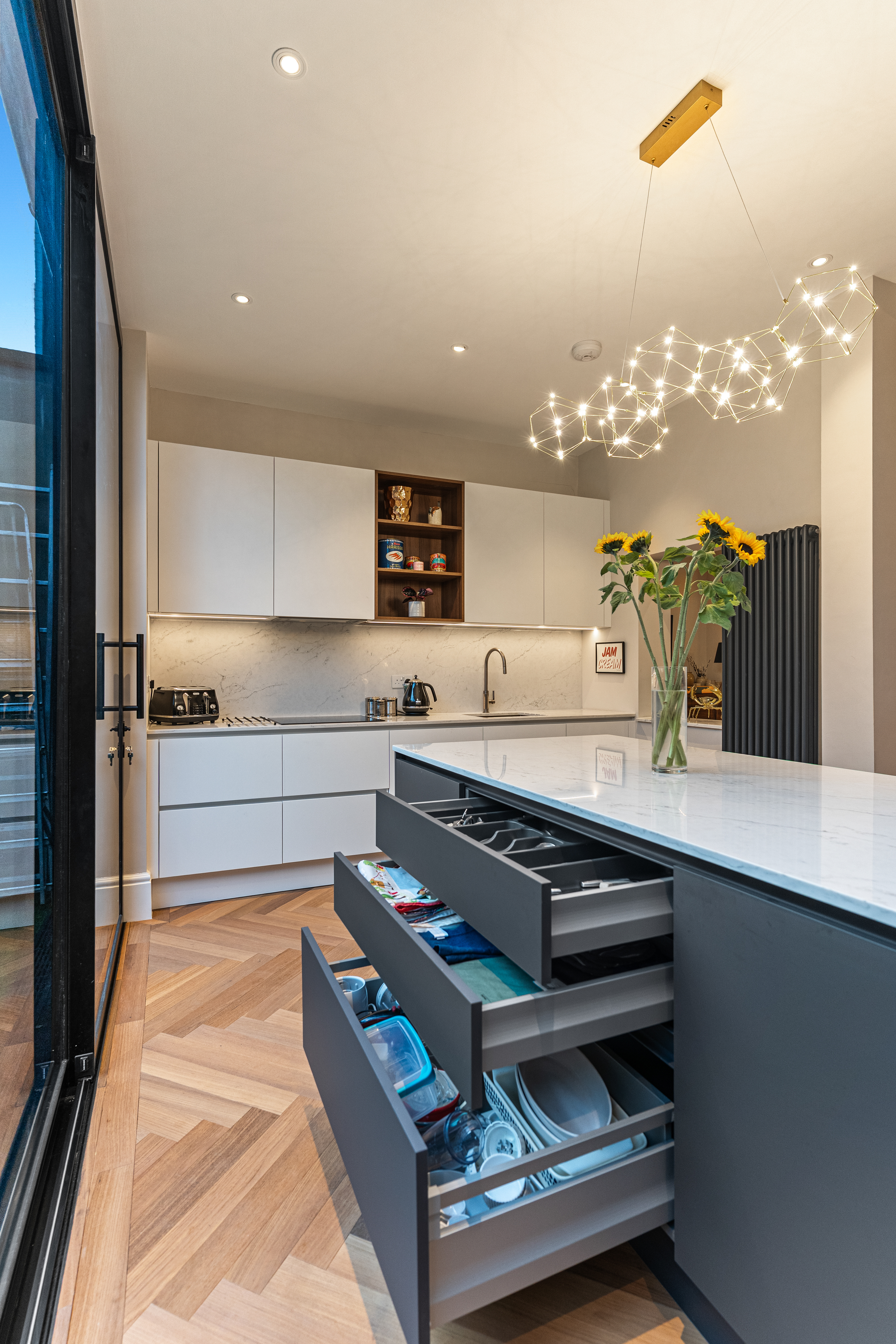 Modern kitchen interior featuring a sleek design with white cabinetry, a marble countertop, and stylish pendant lighting. The image showcases an Arch KBB team member near a kitchen island with open drawers displaying kitchenware and utensils. Sunflowers in a vase add a touch of colour to the space, complemented by wooden flooring and contemporary appliances. Ideal for showcasing modern kitchen design and functionality.