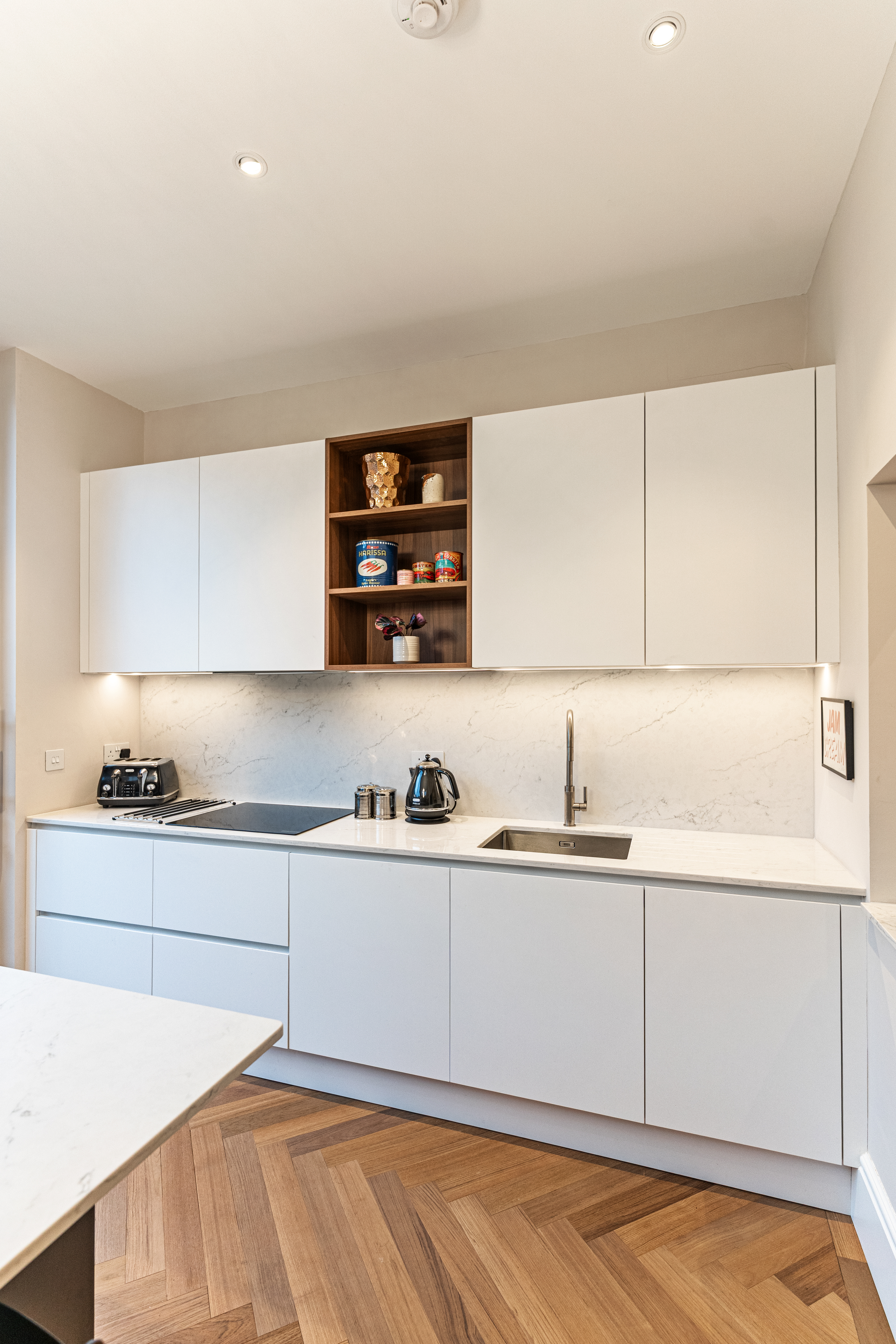 Modern kitchen design featuring sleek white cabinetry, marble countertops, and a wood floor. The space includes a stainless steel sink, a kettle, and a variety of kitchen accessories. An Arch KBB team member is present, showcasing the functionality of the kitchen.
