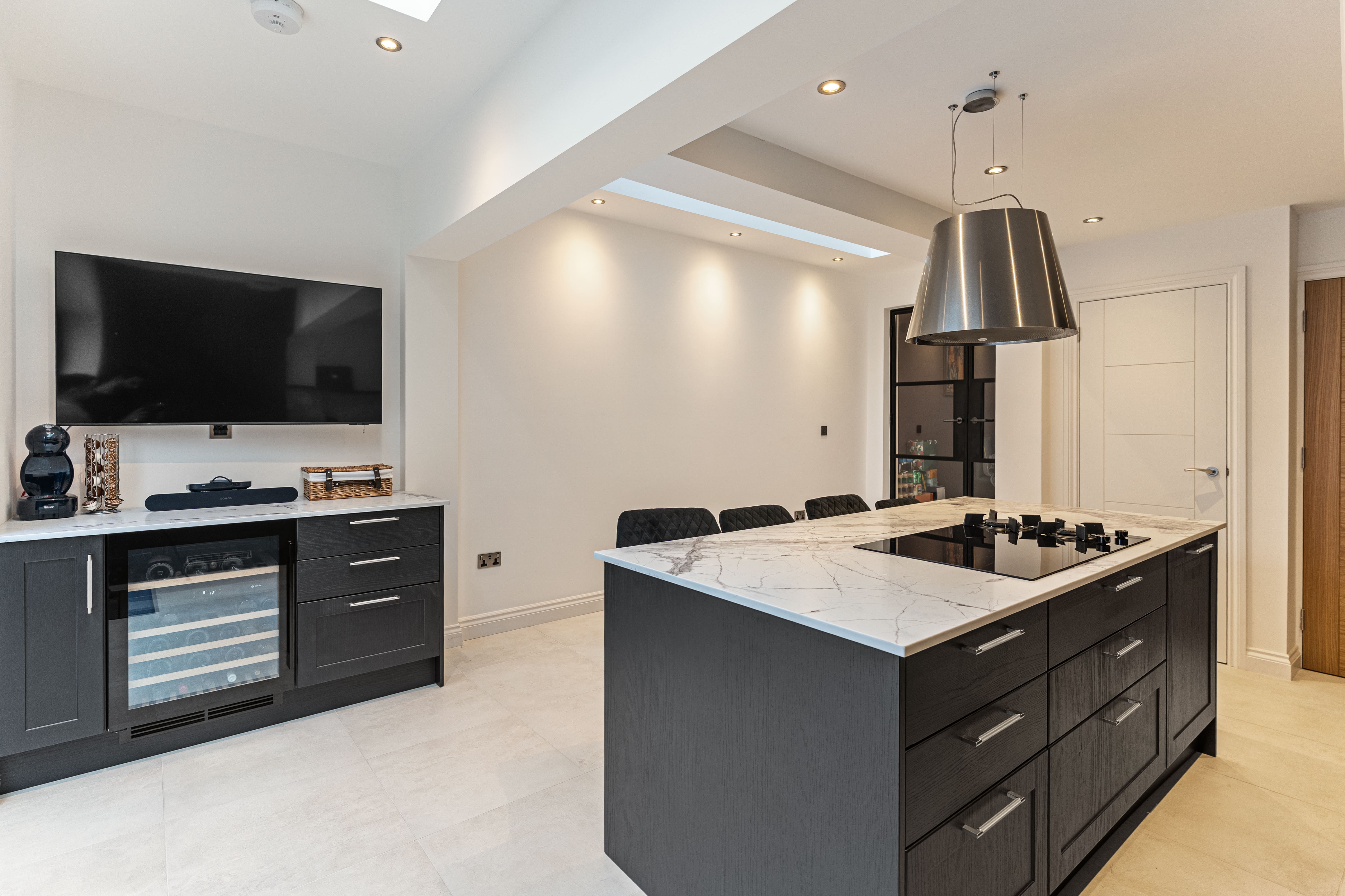 Modern kitchen featuring a stylish island with a marble countertop, sleek black cabinetry, and a built-in wine cooler. A large flat-screen television is mounted on the wall, complemented by contemporary lighting fixtures and a dining area with upholstered chairs. An Arch KBB team member is present, showcasing the kitchen's design and functionality.