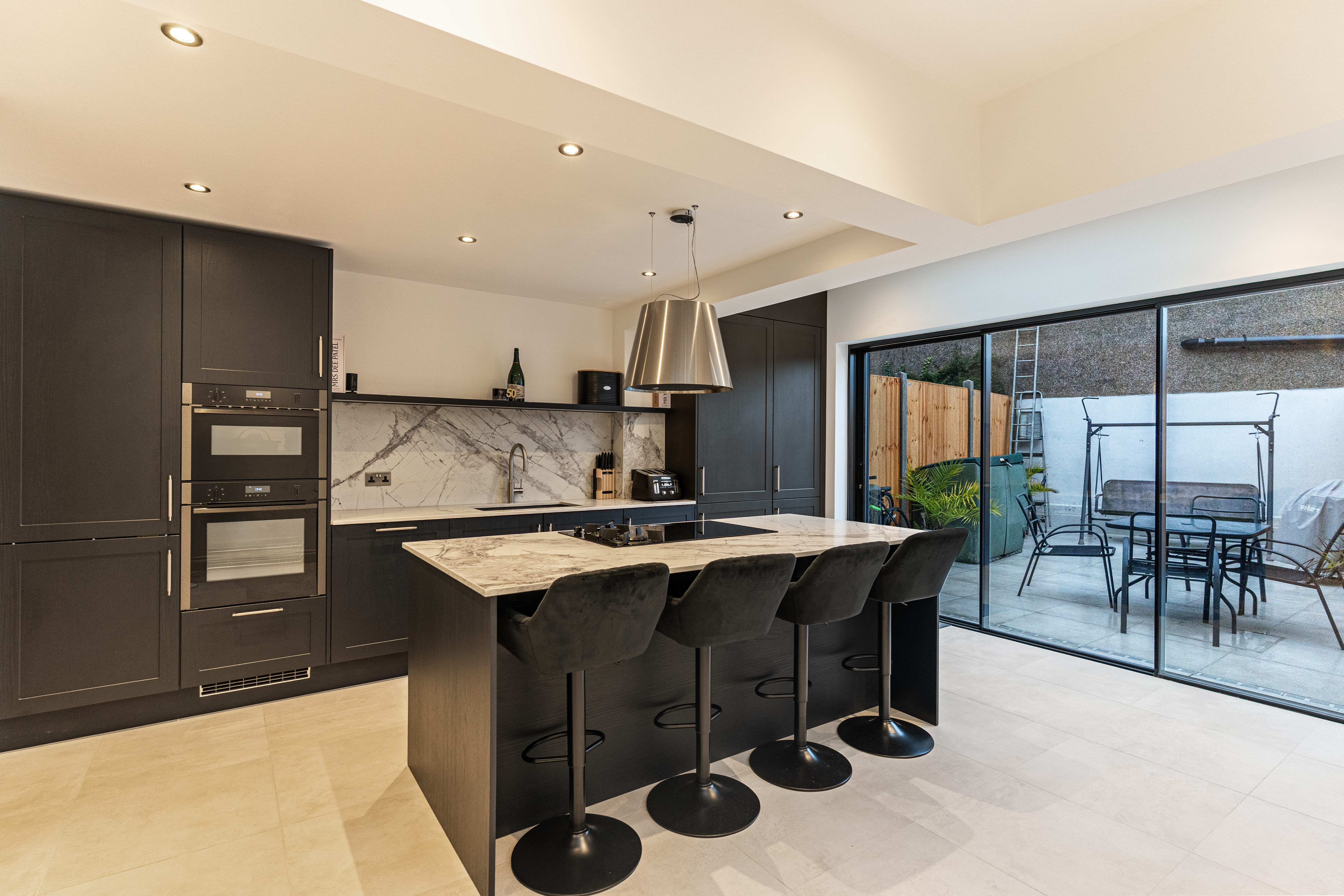 Modern kitchen featuring sleek black cabinetry, a marble-topped island with bar stools, and built-in appliances. Large glass doors provide a view of the outdoor seating area, enhancing natural light and connectivity with the garden. An Arch KBB team member is present, showcasing the kitchen's design.