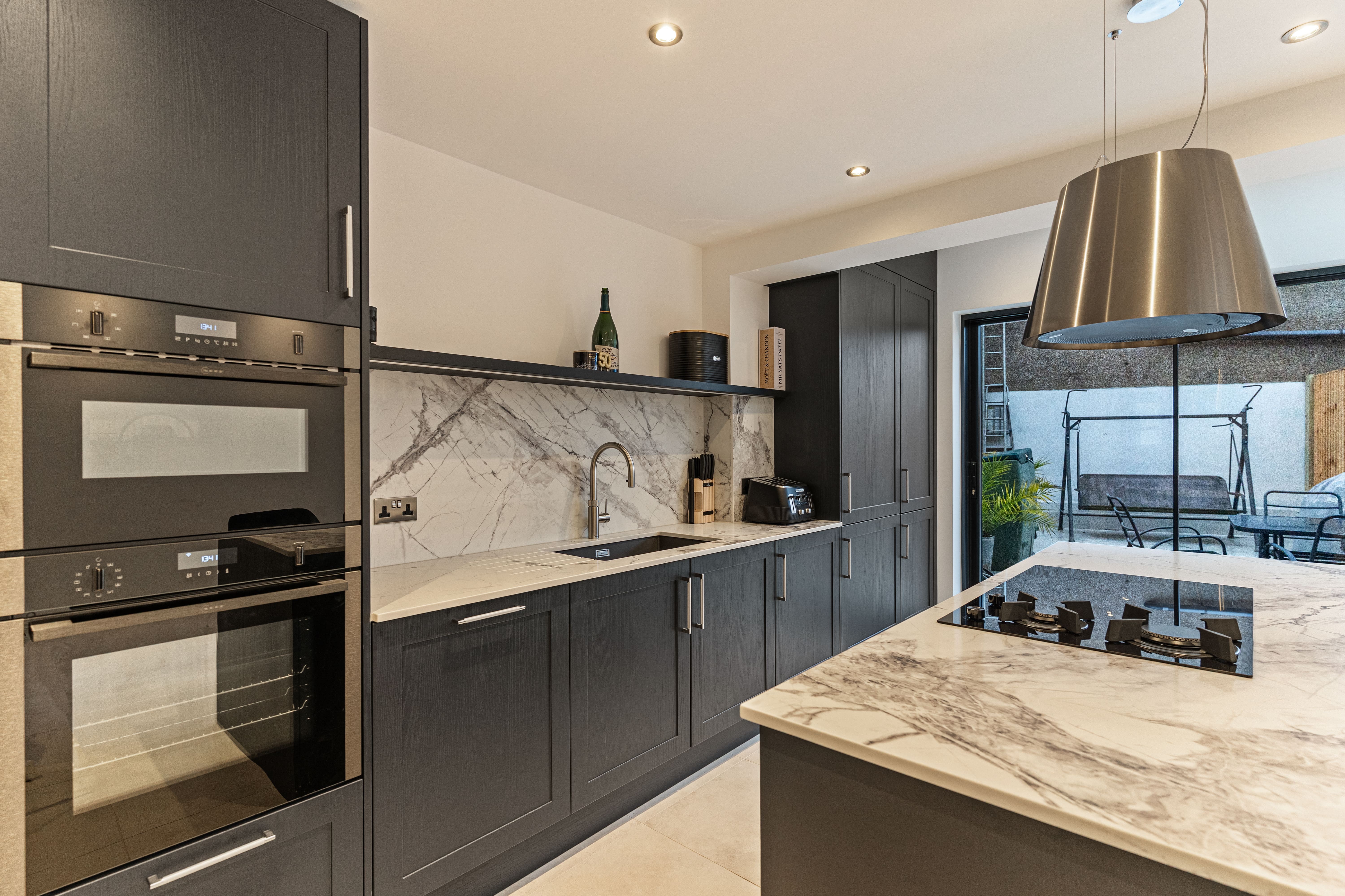 Modern kitchen interior featuring sleek black cabinetry, integrated appliances, and a stylish marble countertop. The design includes a contemporary hanging light fixture and a view of an outdoor seating area through large glass doors. An Arch KBB team member is present, showcasing the kitchen's functionality and aesthetic appeal.
