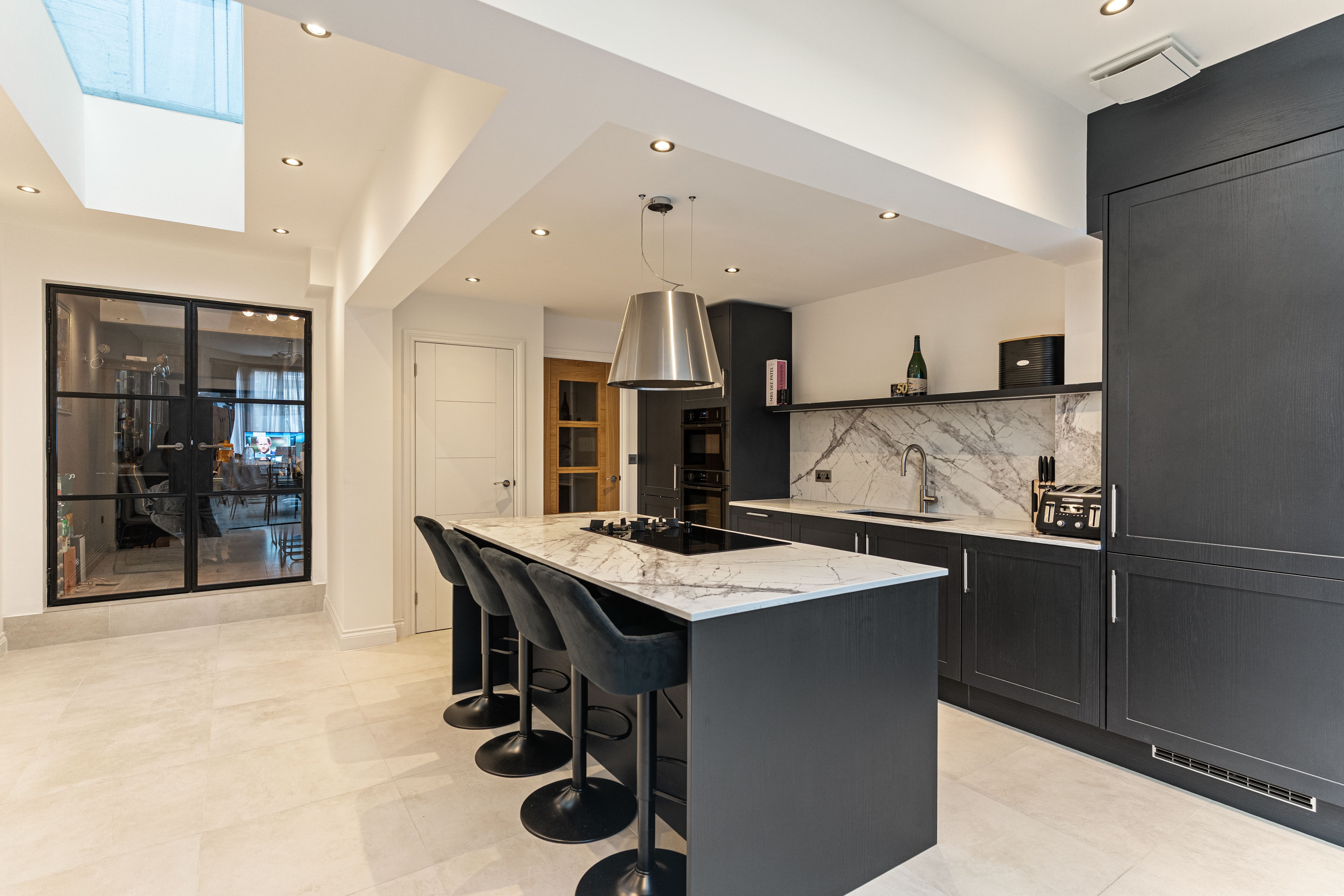 Modern kitchen design featuring a large marble island with black bar stools, sleek cabinetry, and a stylish ceiling light fixture. An Arch KBB team member is visible, showcasing the space's contemporary aesthetic and functionality. Natural light floods the area through a skylight, enhancing the inviting atmosphere.