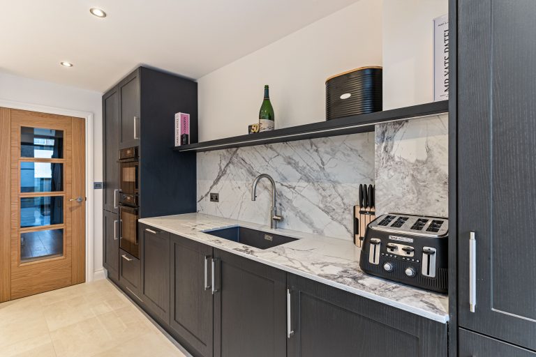 Modern kitchen interior featuring dark cabinetry, marble backsplash, and stainless steel fixtures. Includes a sleek sink, a toaster, and a decorative display on the shelf. An Arch KBB team member is present, showcasing the design.