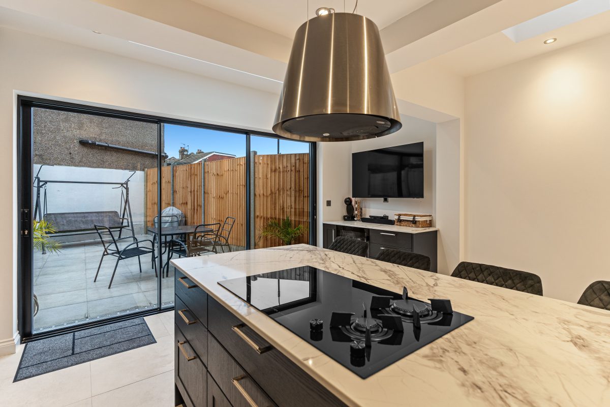 Modern kitchen interior featuring a sleek black gas hob on a marble countertop, with a view of a patio through large sliding doors. An Arch KBB team member is present, showcasing the contemporary design and functionality of the space, which includes a television and stylish cabinetry.