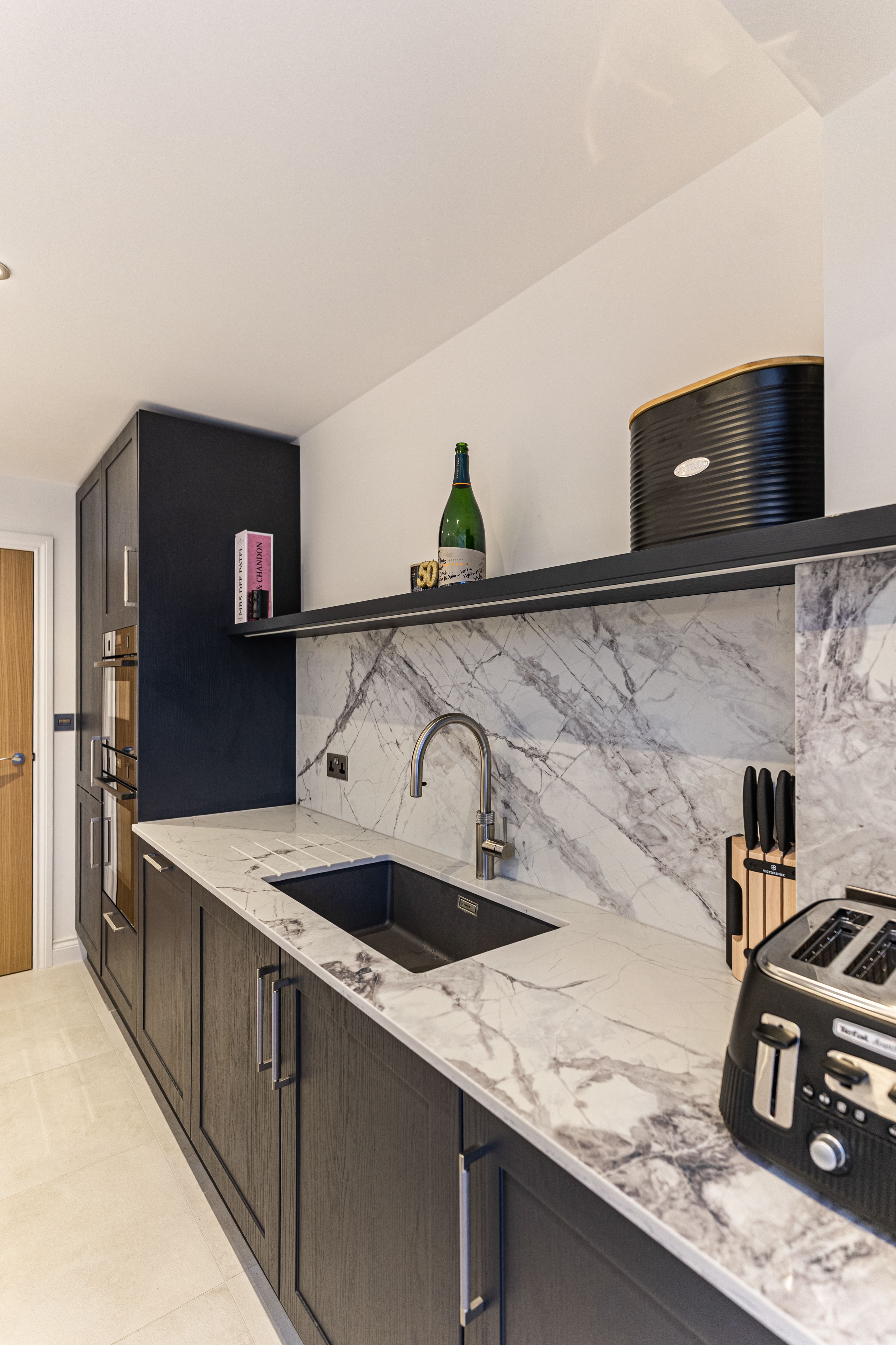 Modern kitchen featuring sleek dark cabinetry, a marble countertop with an integrated sink, and stylish appliances. Decorated with a bottle of champagne and kitchen utensils, this space exemplifies contemporary design. An Arch KBB team member is present, showcasing the kitchen's functionality.
