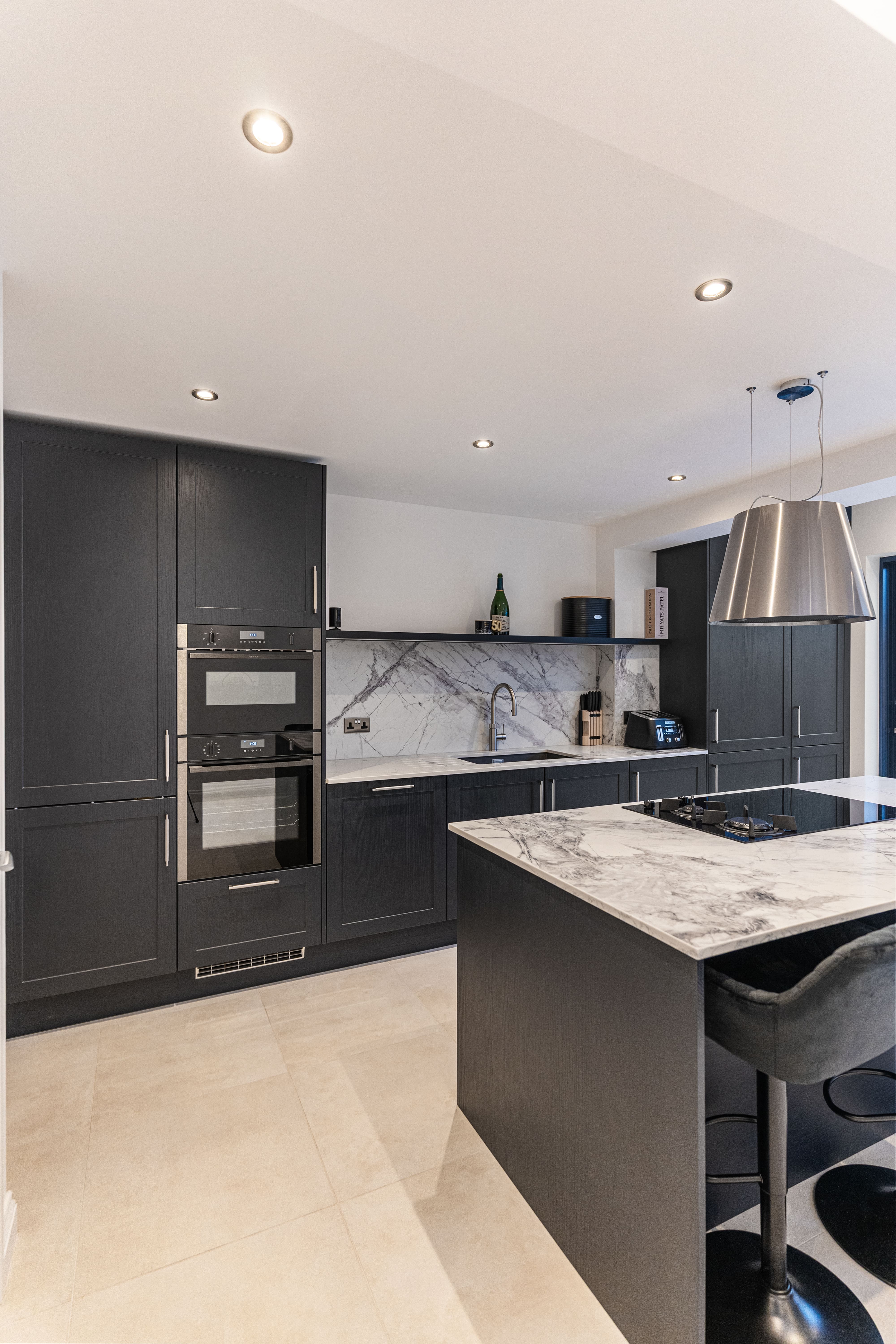 Modern kitchen featuring sleek dark cabinetry, a marble countertop, and integrated appliances. The design includes a stylish island with seating, pendant lighting, and a contemporary backsplash. An Arch KBB team member is present, showcasing the kitchen's functionality and aesthetic appeal.