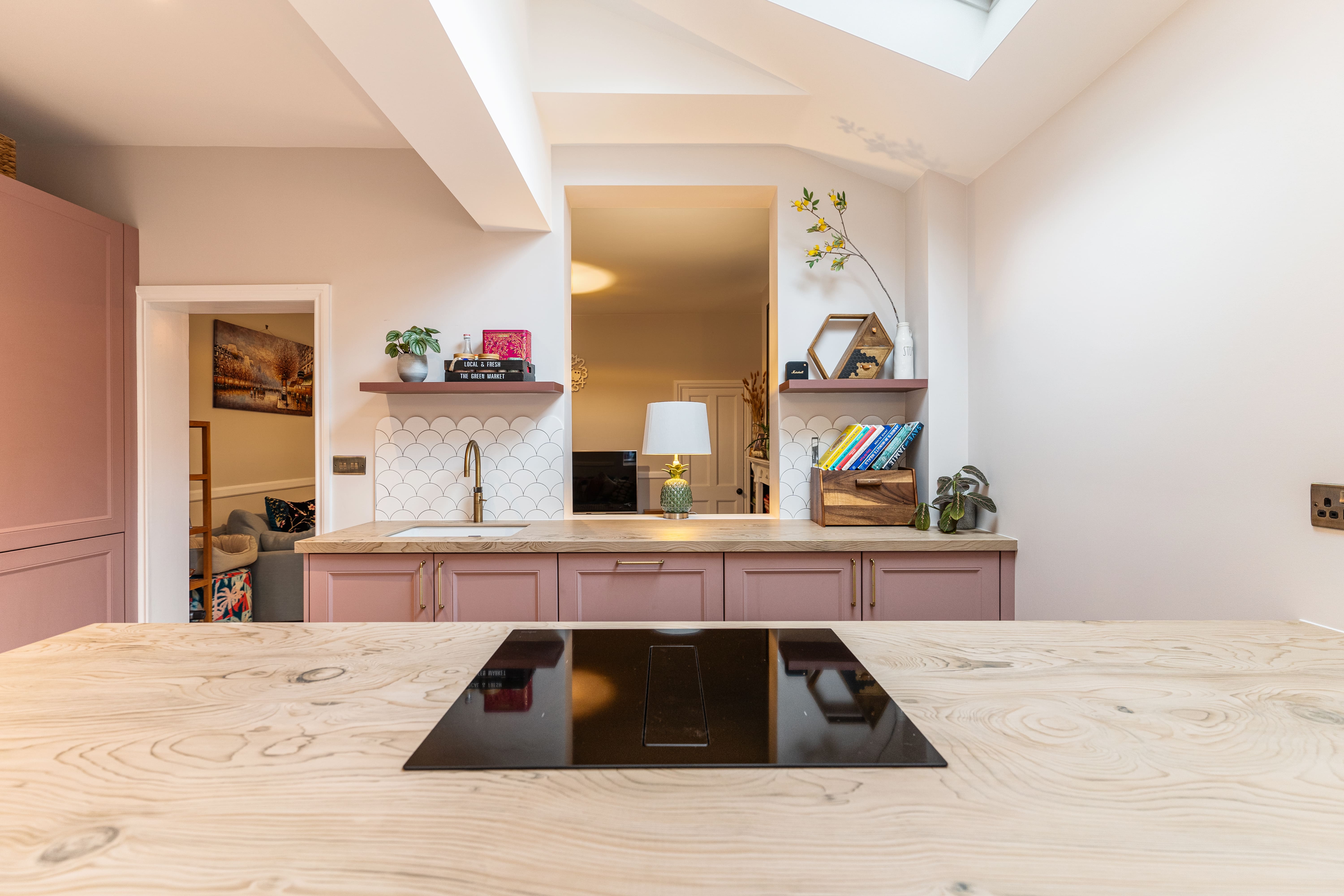 Modern kitchen interior featuring a wooden countertop, a black induction hob, and pink cabinetry. The space includes decorative shelves with books and plants, a stylish lamp, and a tiled backsplash. Natural light floods the area, enhancing the contemporary design. An Arch KBB team member is present, contributing to the kitchen's aesthetic appeal.