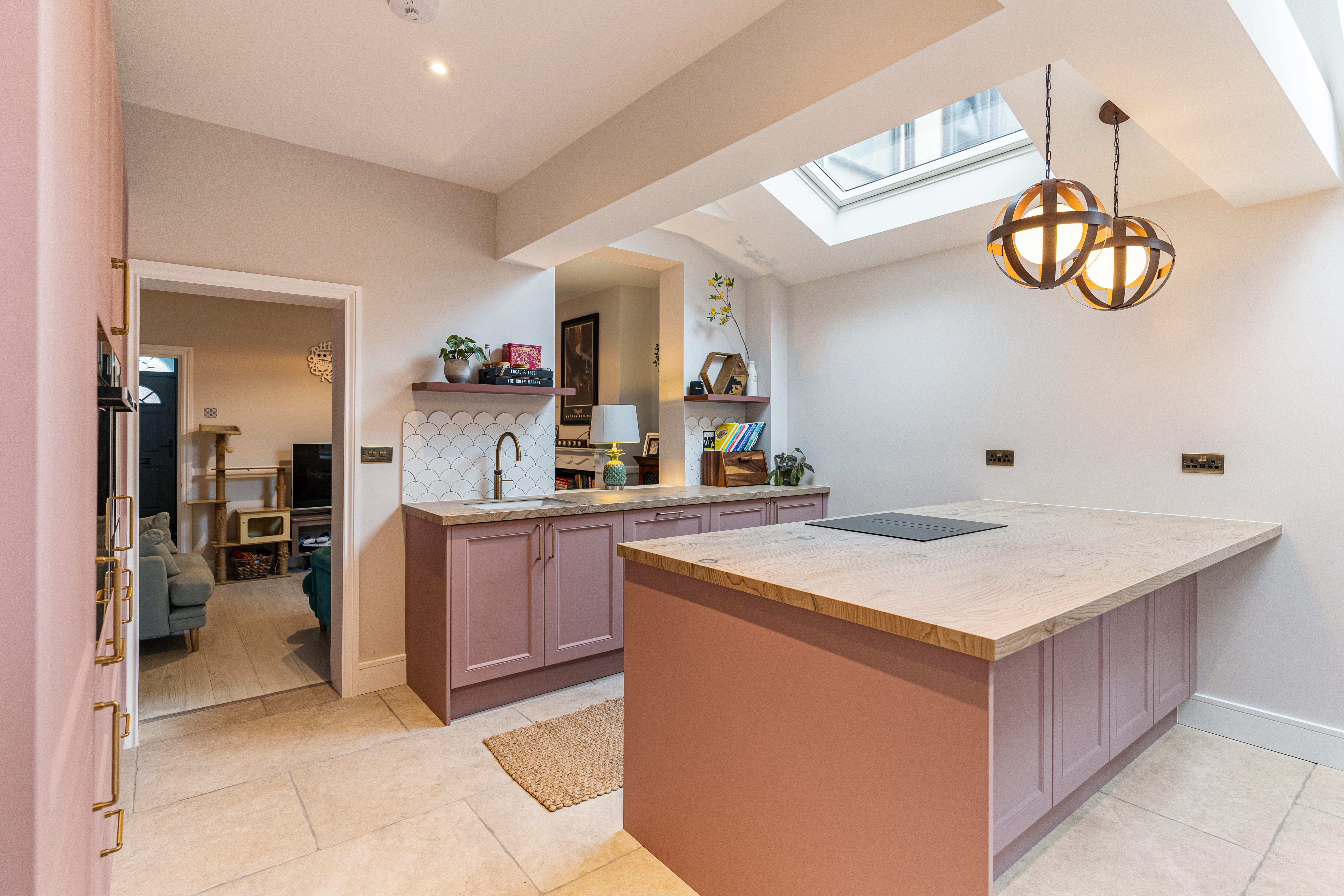 Modern kitchen featuring a spacious layout with pink cabinetry and a wooden countertop. The room is illuminated by stylish pendant lights and includes a skylight for natural light. An Arch KBB team member is present, showcasing the kitchen's design elements, which include a patterned backsplash and open shelving for decorative items.