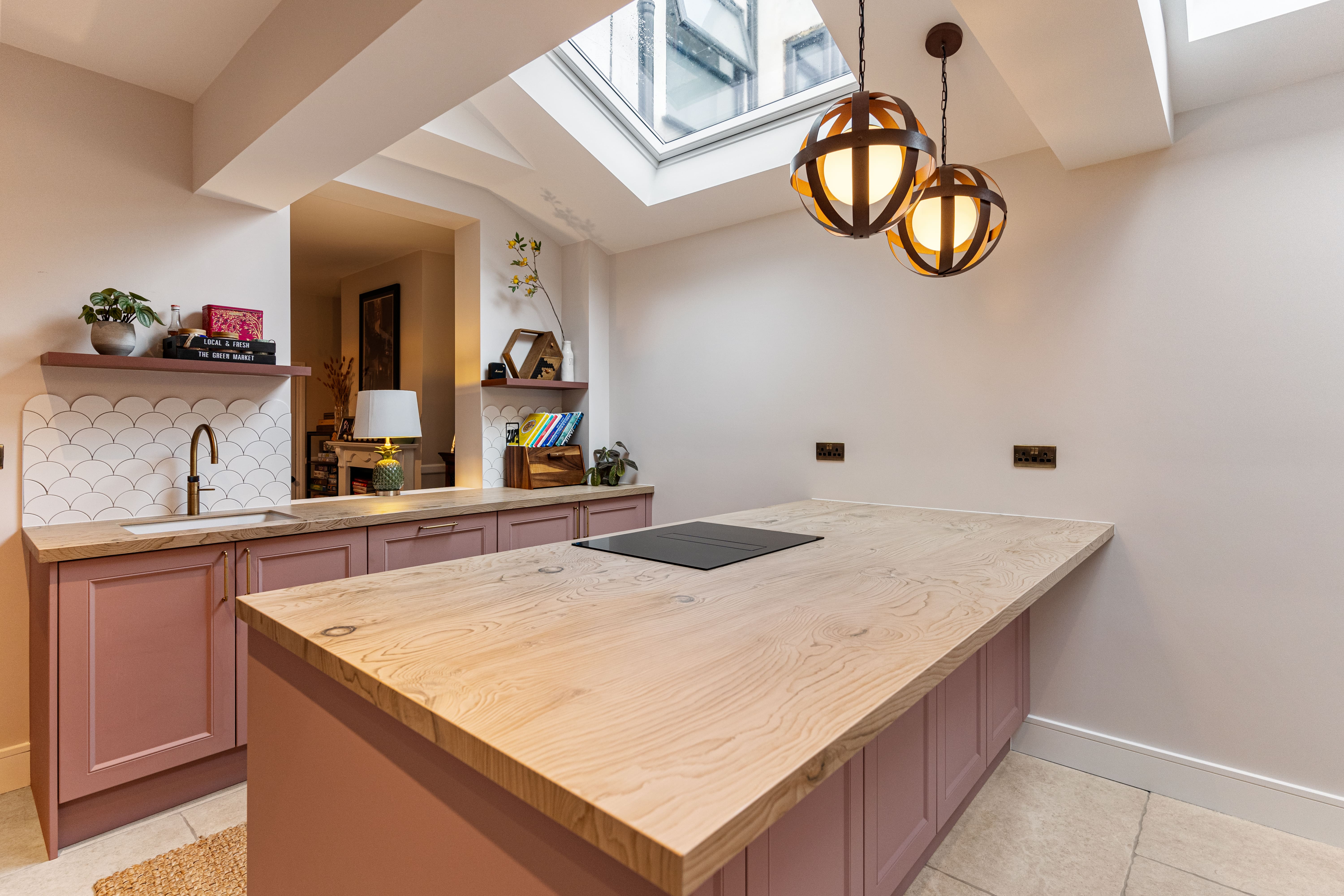 Modern kitchen interior featuring a large wooden island with an integrated hob, pastel pink cabinetry, and a tiled splashback. The space is illuminated by stylish pendant lights and natural light from a skylight, creating a warm and inviting atmosphere. An Arch KBB team member is present, showcasing the design.