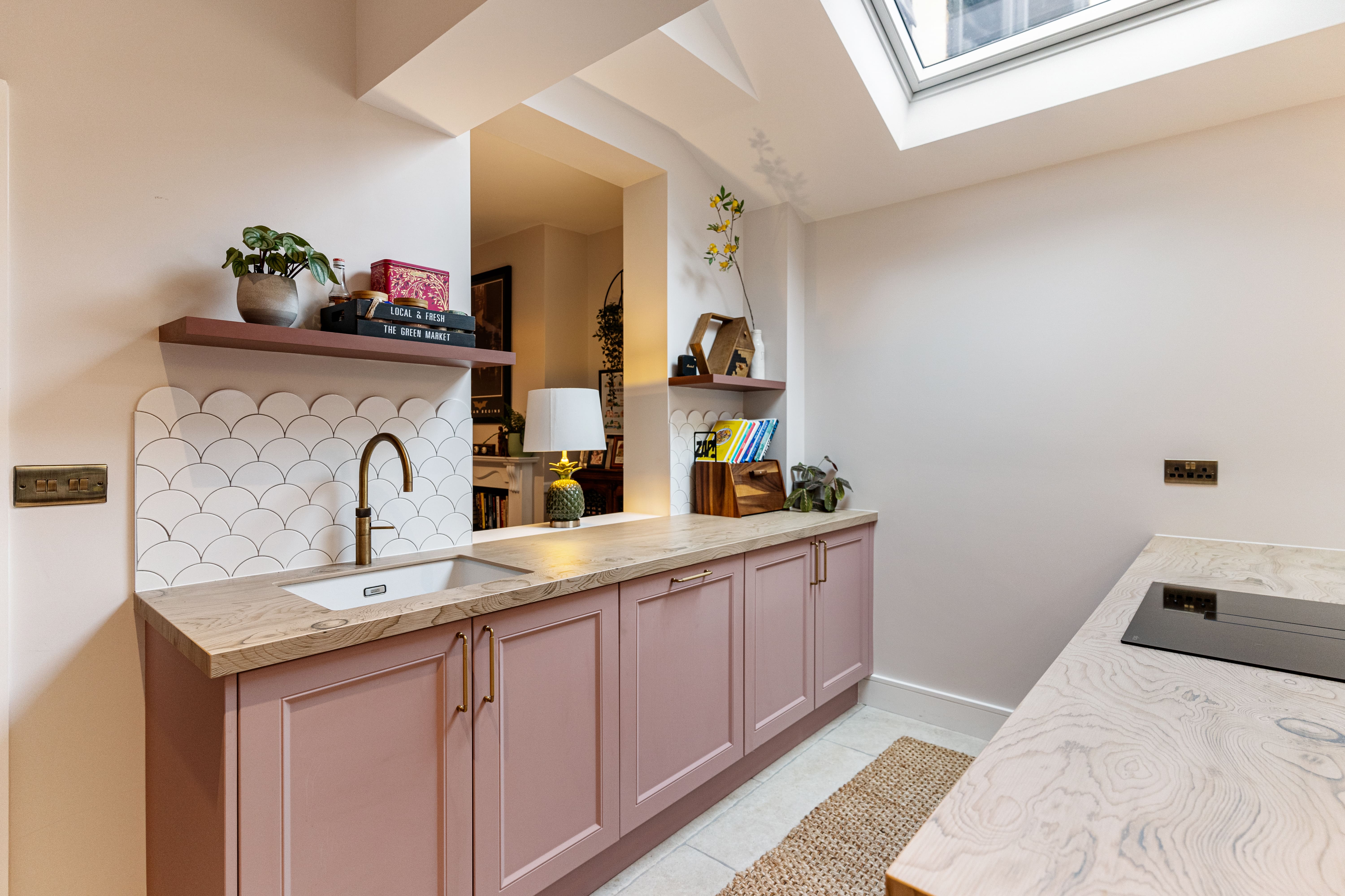 Modern kitchen design featuring a pink cabinetry, a stylish sink with a gold tap, and a decorative tiled backsplash. Open shelving displays plants, books, and decorative items, while natural light floods the space through a skylight. An Arch KBB team member is present, contributing to the design atmosphere.