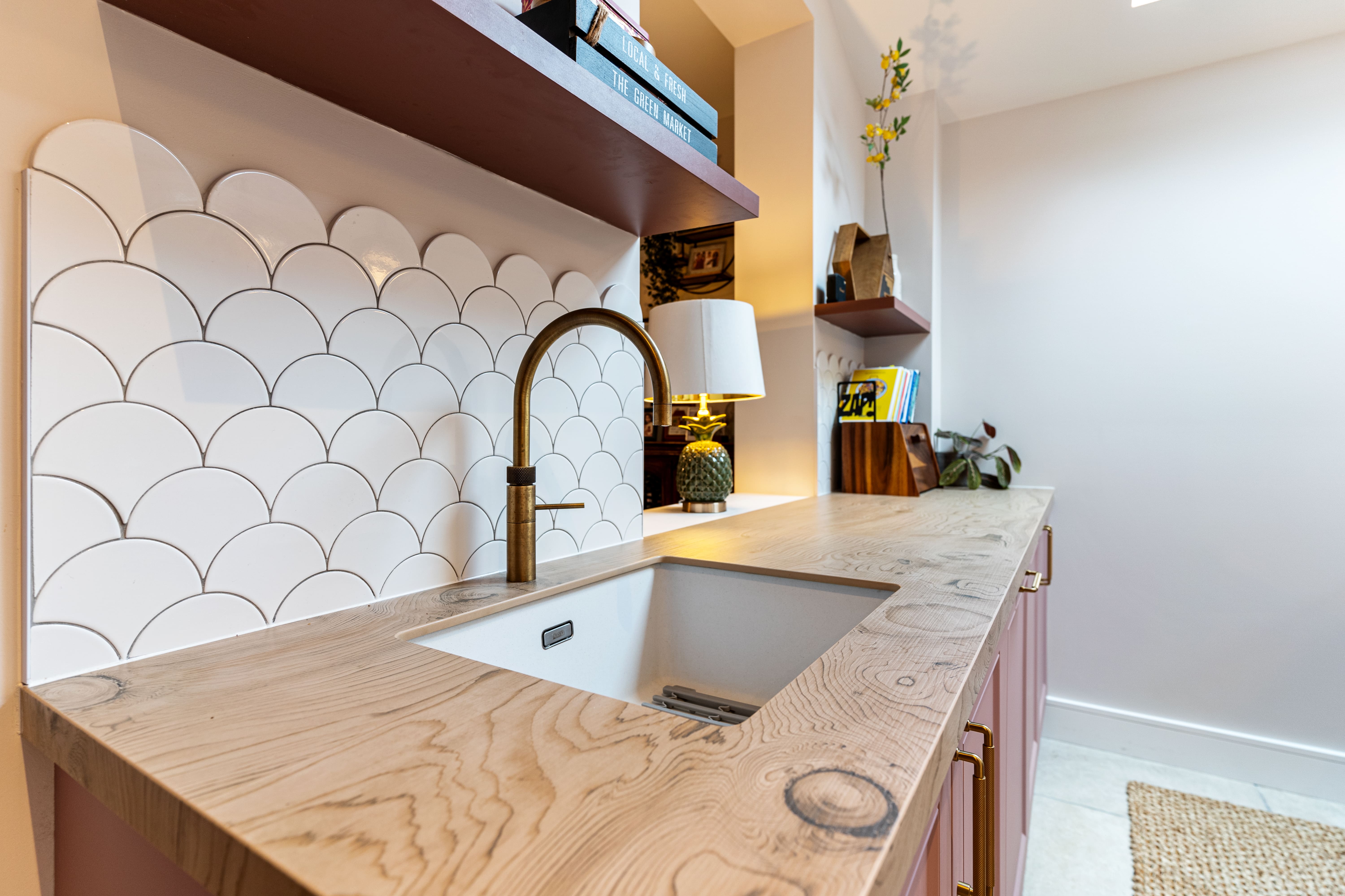 A modern kitchen featuring a wooden countertop with a built-in sink, complemented by a stylish brass tap. The backsplash showcases white scalloped tiles, while open shelves above display decorative items and books. An Arch KBB team member is present, enhancing the inviting atmosphere of the space.