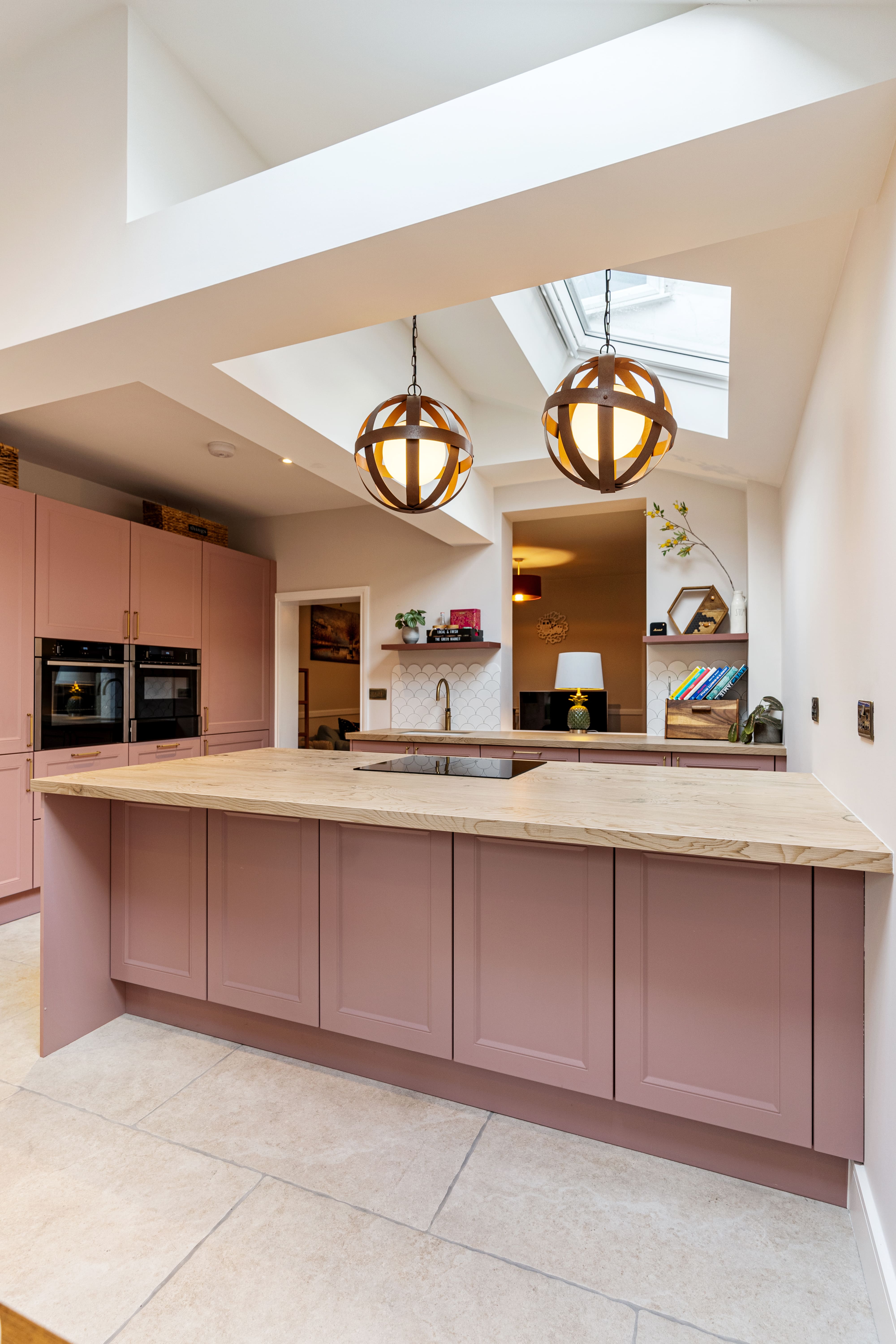 Modern kitchen design featuring a spacious island with a wooden countertop and pink cabinetry. The room is illuminated by stylish pendant lights and has a skylight that enhances natural light. An Arch KBB team member is present, highlighting the inviting atmosphere of the space.