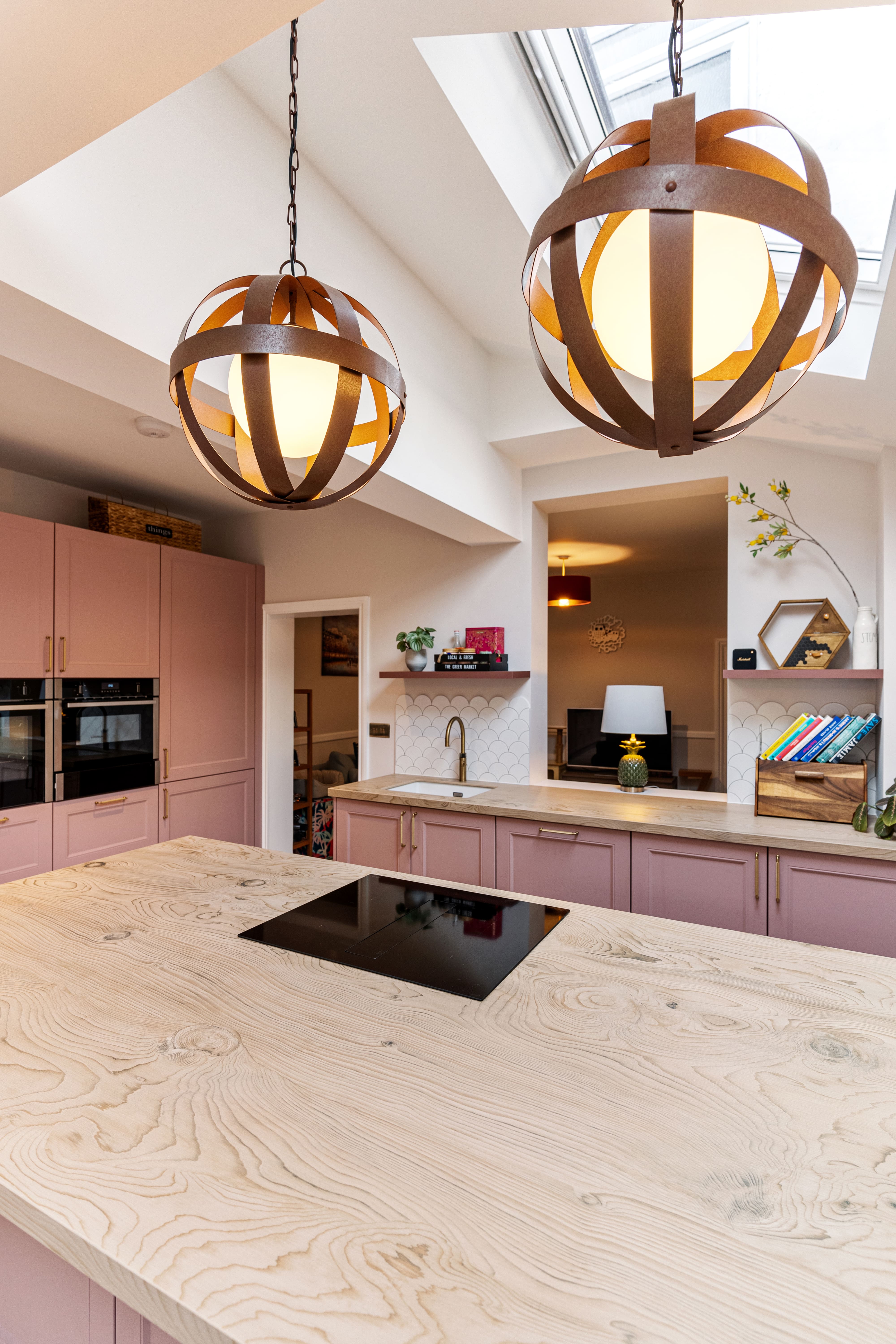 Modern kitchen interior featuring pink cabinetry, a wooden island countertop, and black induction hob. Two stylish pendant lights hang above the island, illuminating the space. An Arch KBB team member is present, showcasing a well-designed and functional kitchen environment.