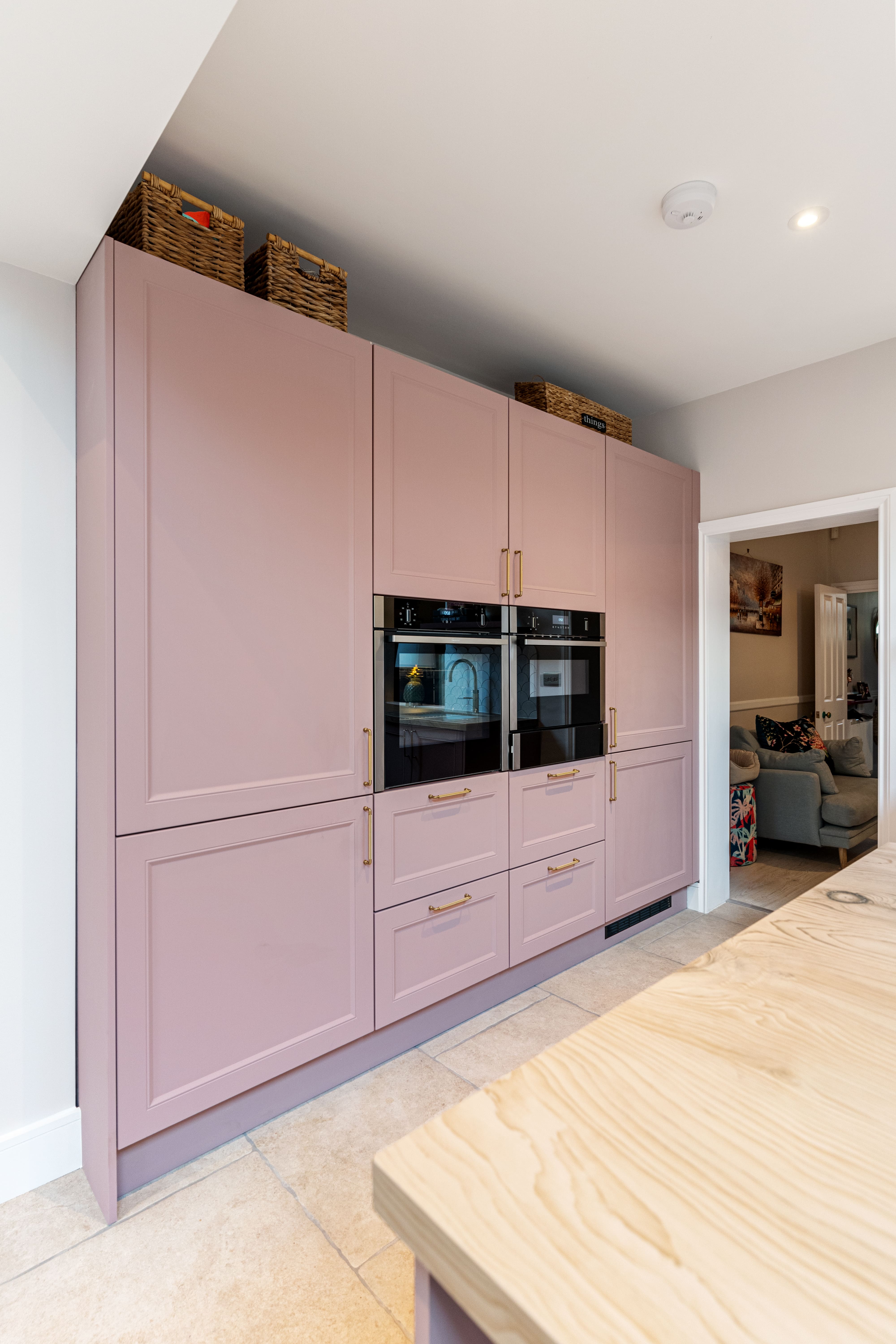 Modern kitchen design featuring a pink cabinetry with gold handles, integrated black ovens, and a wooden worktop. The space is well-lit, showcasing an organised aesthetic with decorative baskets atop the cabinets. An Arch KBB team member is present, contributing to the kitchen's stylish interior.