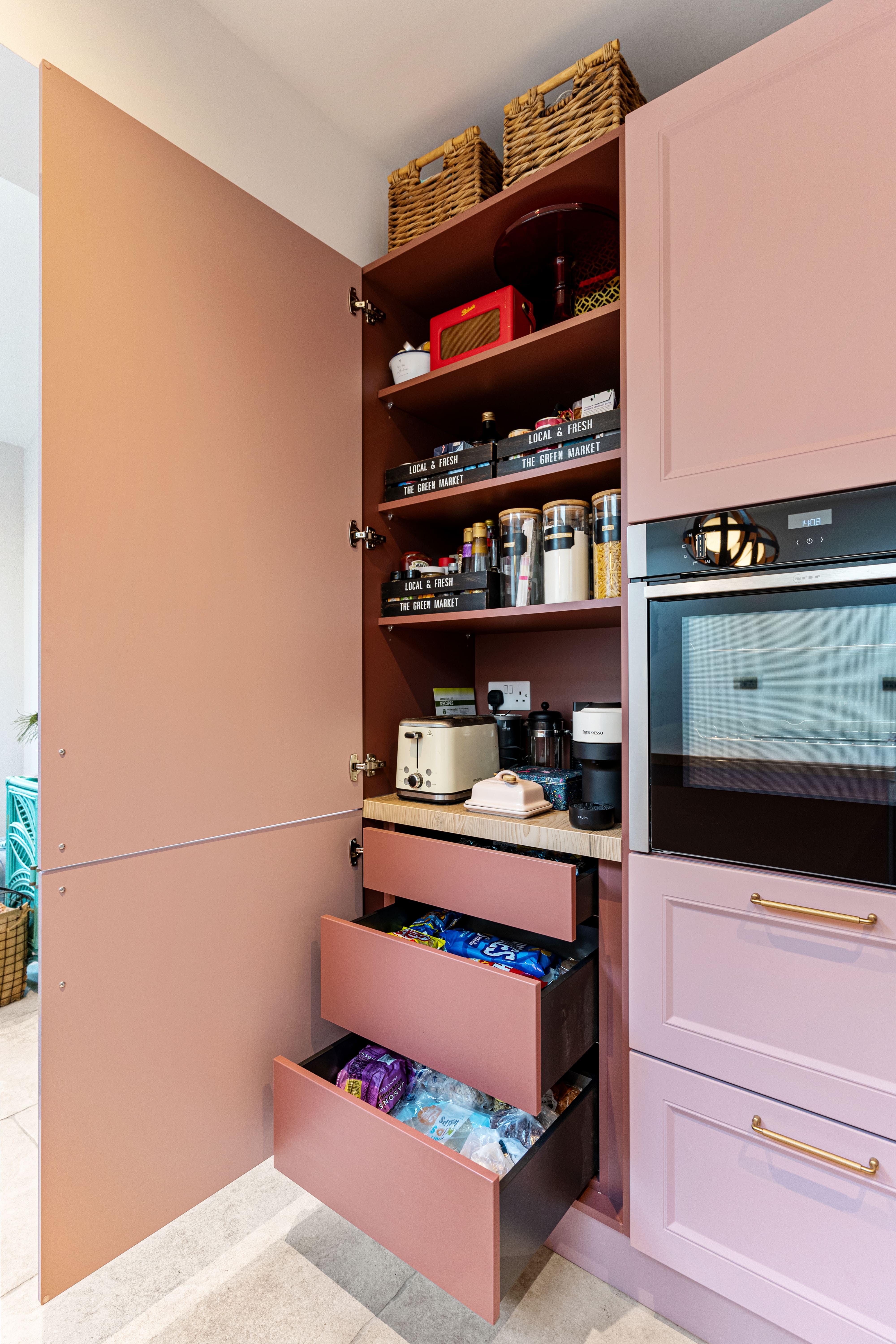 Interior view of a stylish kitchen pantry featuring pink cabinetry, neatly organised shelves with various jars and containers, a toaster, and coffee maker. The pantry includes three spacious drawers filled with snacks, showcasing efficient storage solutions. An Arch KBB team member is present, contributing to the design.
