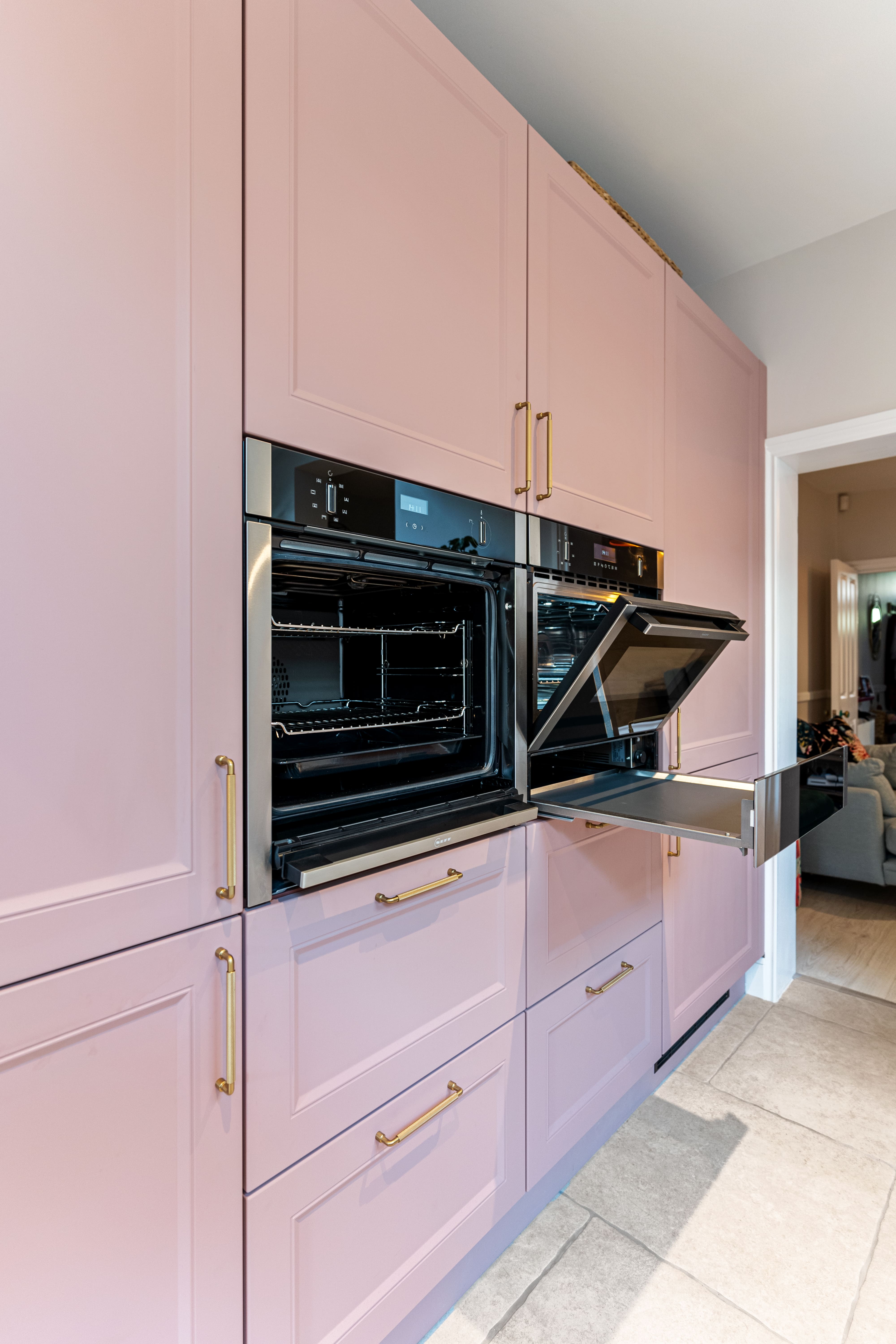 Modern kitchen featuring stylish pink cabinetry with gold handles and two black built-in ovens, one of which is open. An Arch KBB team member is present, showcasing the kitchen's elegant design and functionality.