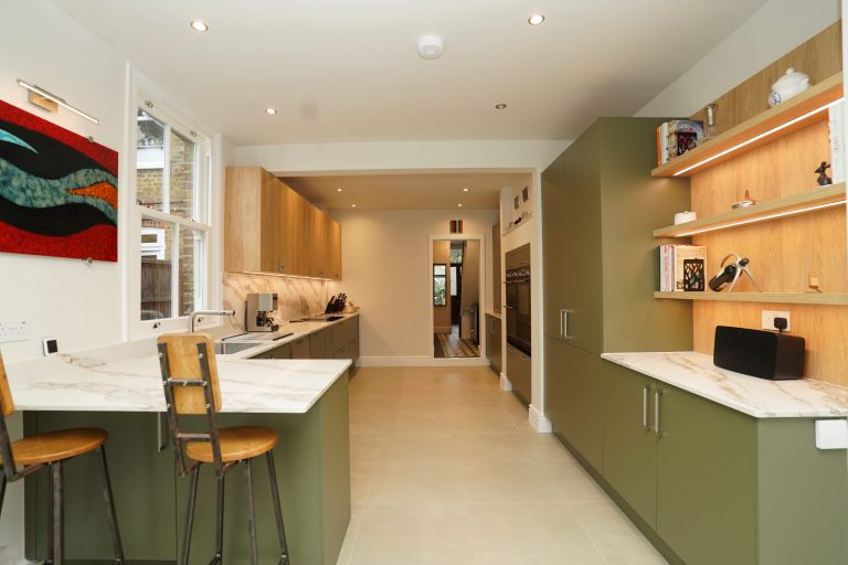 Modern kitchen design by Arch KBB featuring olive green cabinets, marble countertops, and wood shelving. The space includes bar stools, integrated appliances, and contemporary artwork, creating a stylish and functional cooking environment.