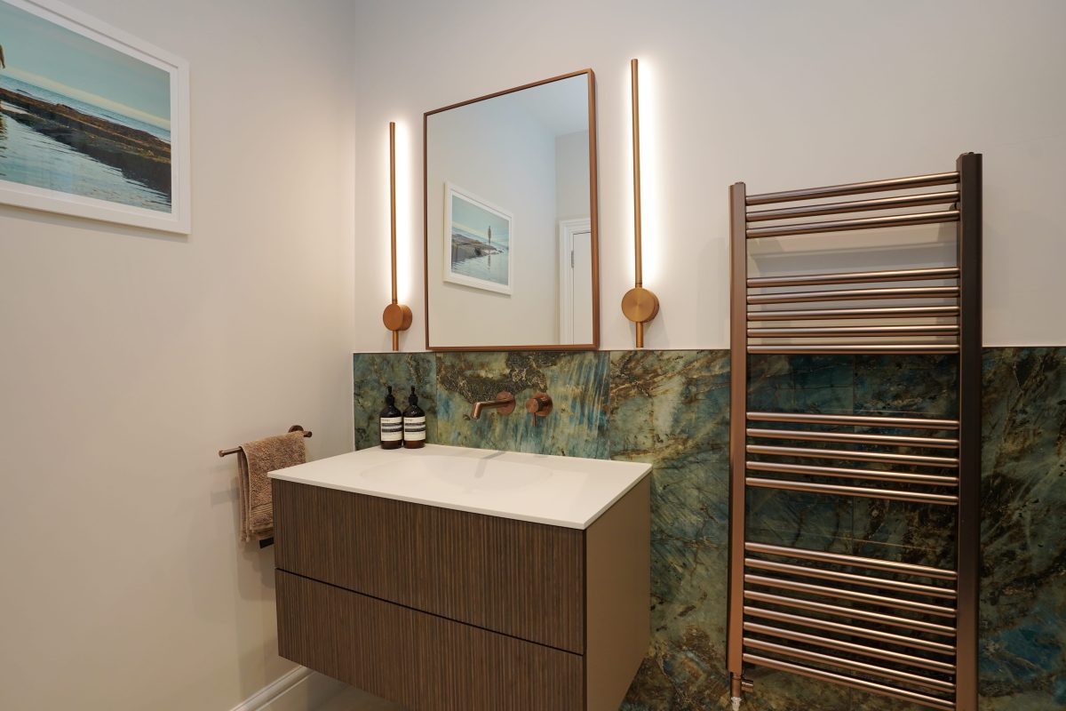 Modern bathroom featuring a stylish vanity with a white countertop, illuminated mirror, and a striking green and gold marble splashback. A sleek towel rail and decorative wall art enhance the contemporary design. Arch KBB team member present in the space, showcasing innovative bathroom solutions.