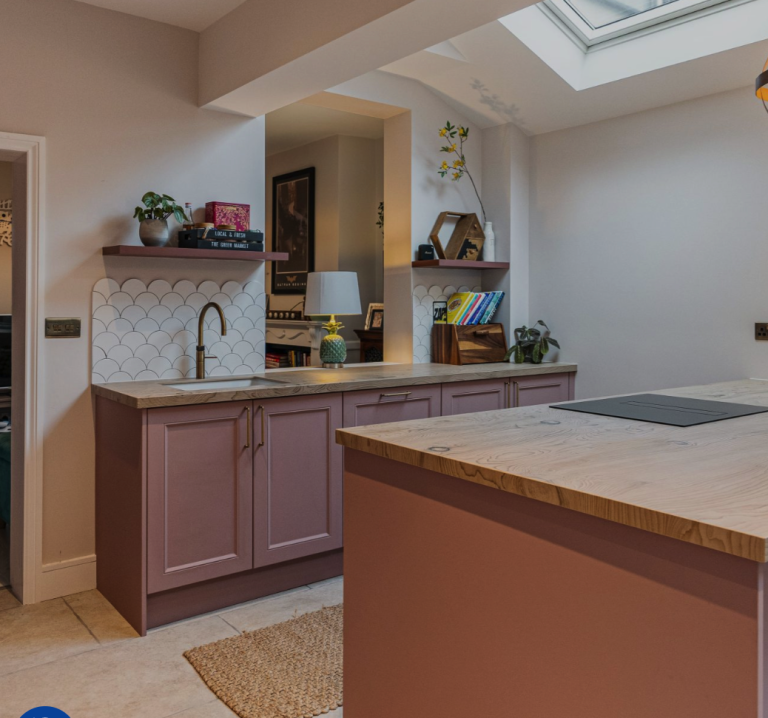 Modern kitchen design featuring a pink cabinetry, wooden worktops, and scalloped tiles. The space is enhanced by natural light from a skylight, with shelves displaying decor and books. An Arch KBB team member is present, contributing to the inviting atmosphere of the kitchen.