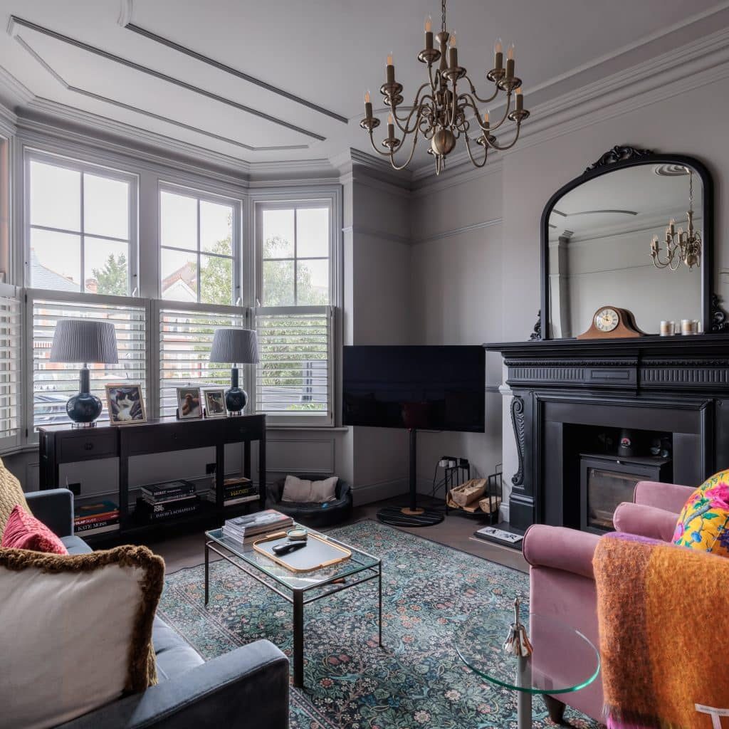 Elegant living room with large bay windows, a classic chandelier, and a fireplace with a decorative mirror. Features plush seating, vibrant cushions, and a patterned rug, creating a cosy and stylish atmosphere.