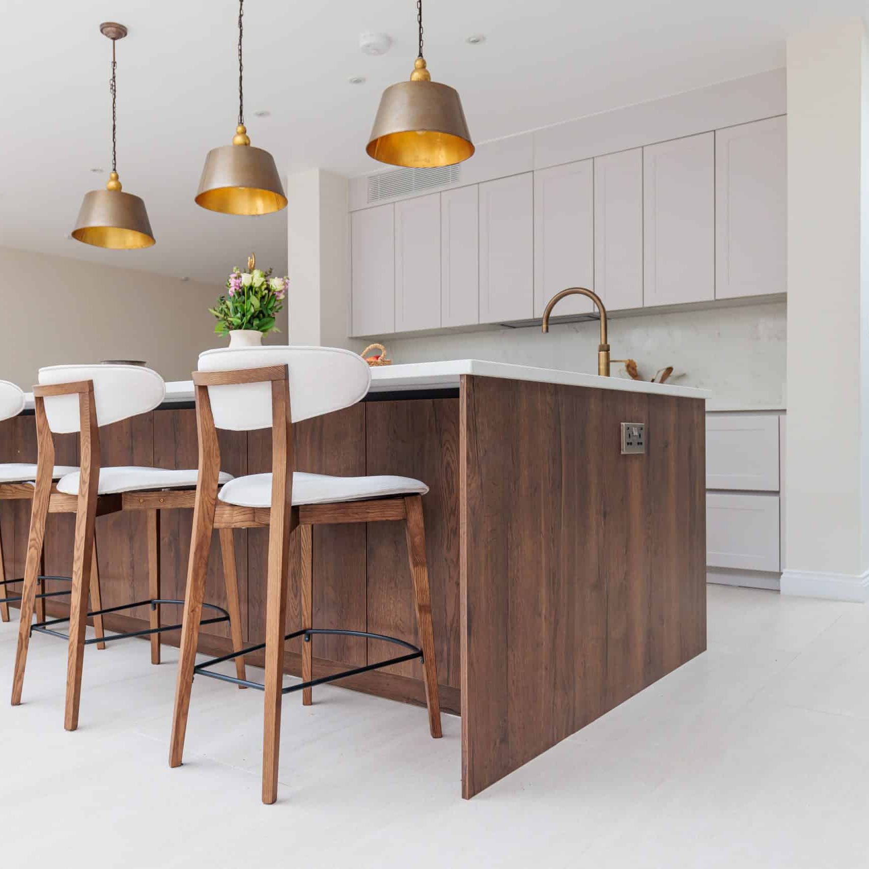 Modern kitchen interior featuring a stylish island with wooden cabinetry and seating for four. Bright pendant lights hang above the island, complementing the contemporary design. The space includes sleek cabinetry and a neutral colour palette, creating an inviting atmosphere. An Arch KBB team member is present, engaging in the kitchen environment.