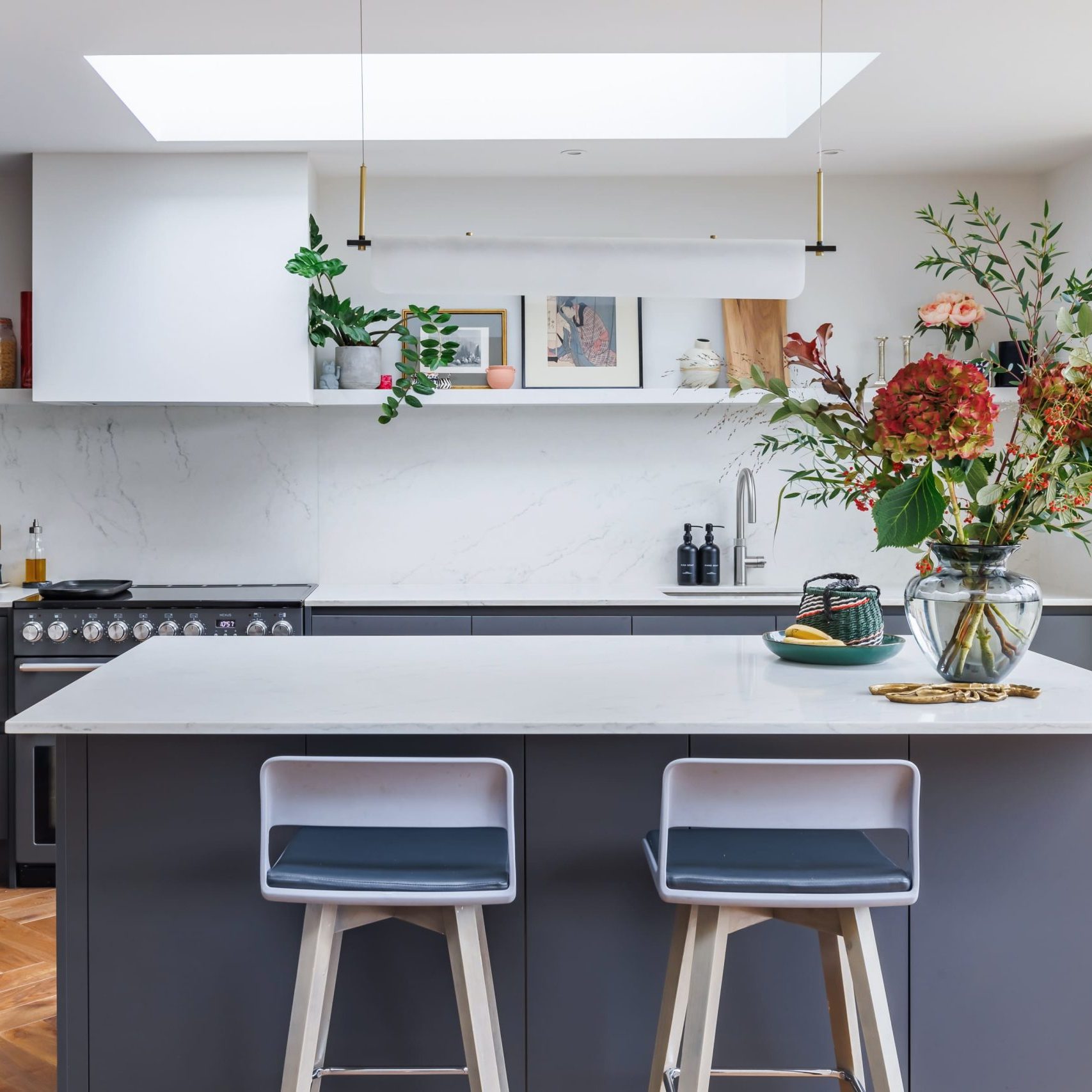 Modern kitchen interior featuring a sleek grey island with two stylish stools, marble countertops, and a skylight above. The space is adorned with vibrant floral arrangements, stylish kitchenware, and tasteful artwork on the walls, creating an inviting and contemporary atmosphere.