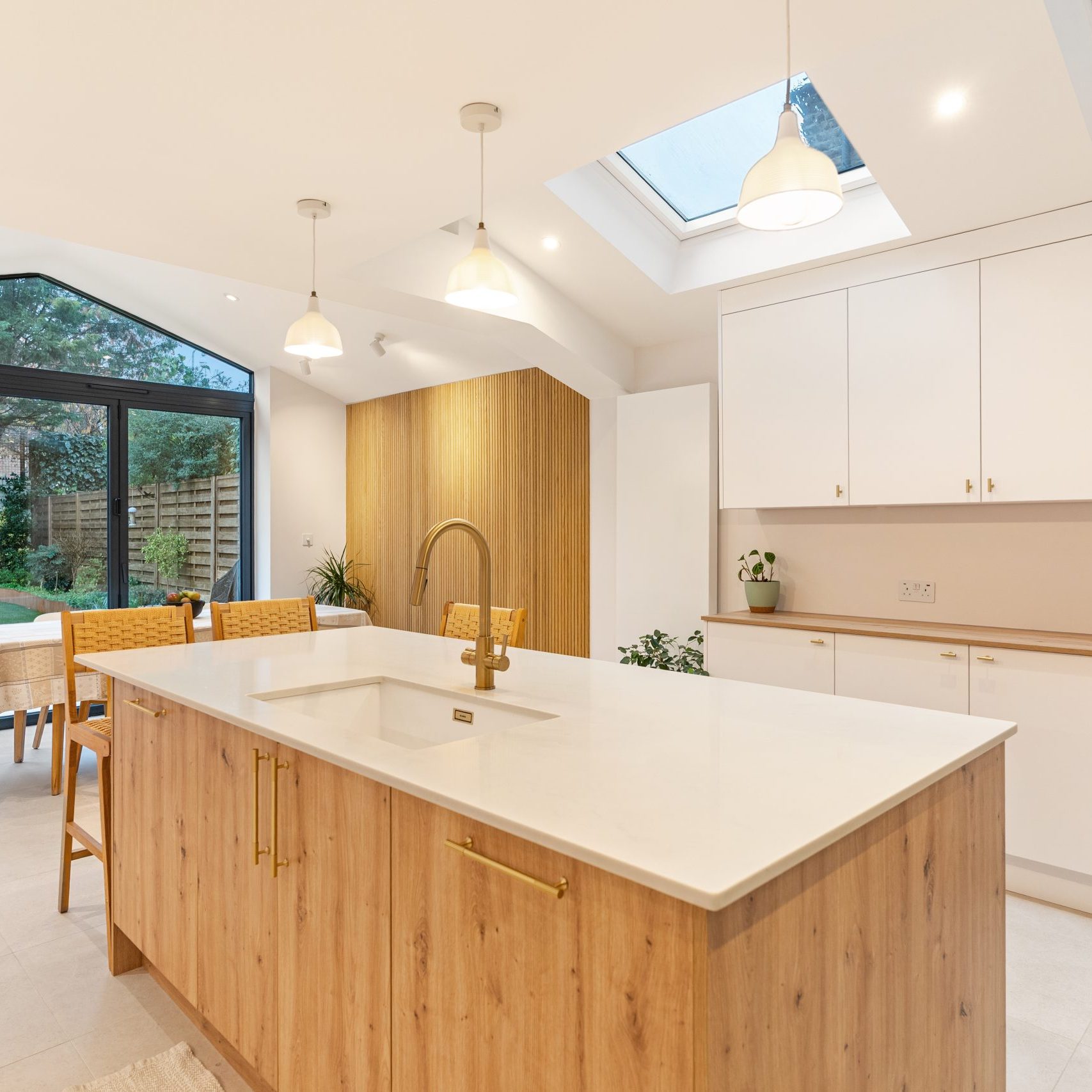 Modern kitchen design featuring an island with a white countertop and wooden cabinetry, complemented by gold fixtures. The space is illuminated by pendant lights and has large glass doors leading to a garden, enhancing natural light and creating an open feel. An Arch KBB team member is present, engaging with the kitchen layout.