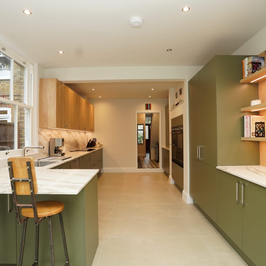 Modern kitchen design by Arch KBB featuring olive green cabinets, marble countertops, and wood shelving. The space includes bar stools, integrated appliances, and contemporary artwork, creating a stylish and functional cooking environment.