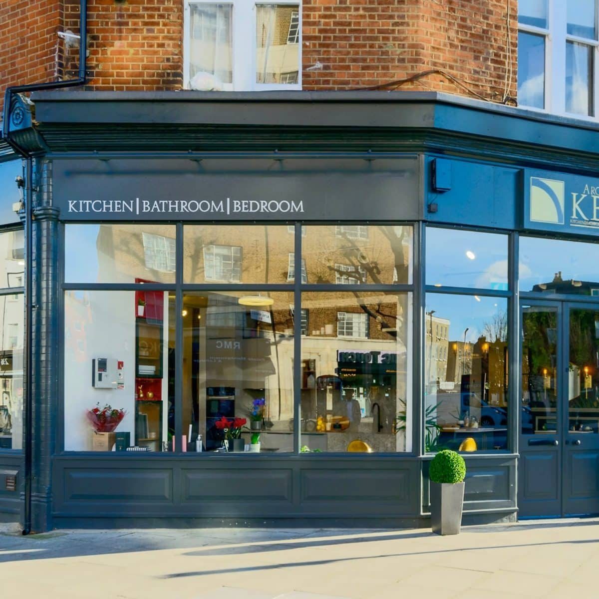 Exterior view of the Arch KBB showroom, showcasing kitchen, bathroom, and bedroom design services, with large glass windows and a brick facade.